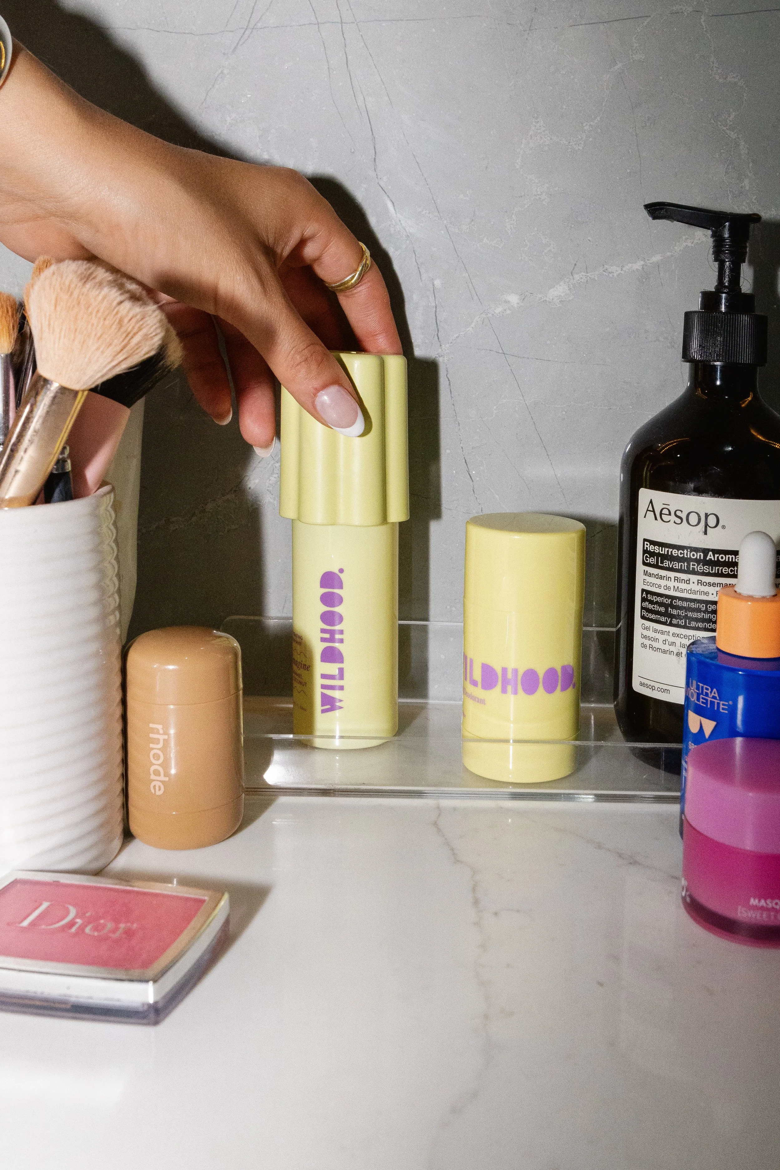 A person's hand reaching for a yellow Wildhood lip balm on a bathroom shelf, surrounded by skincare products, brushes, and a Dior compact.