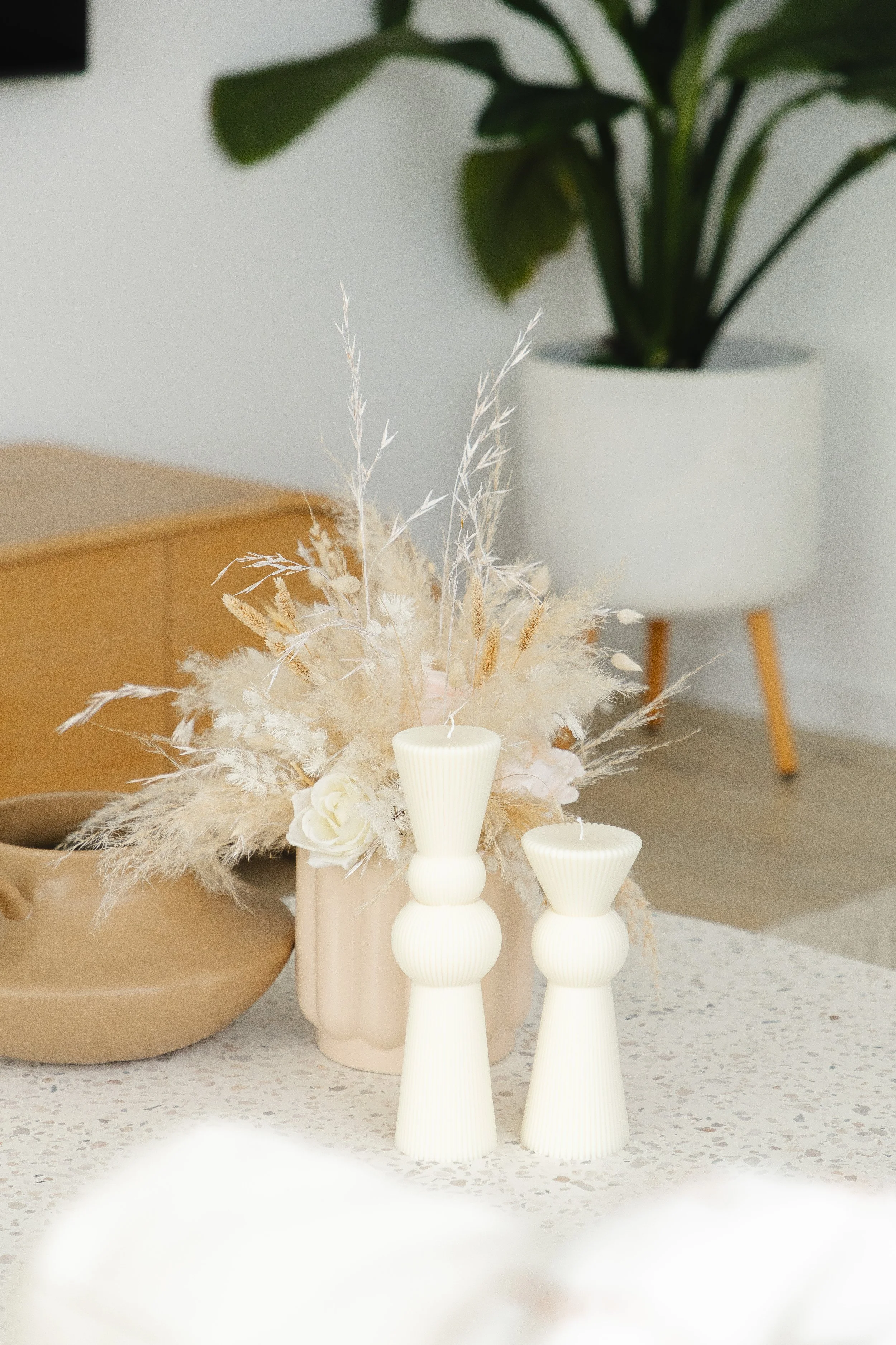 Decorative arrangement with two cream-colored ribbed vases and dried beige and white flowers on a speckled countertop, with a potted green plant in the background.