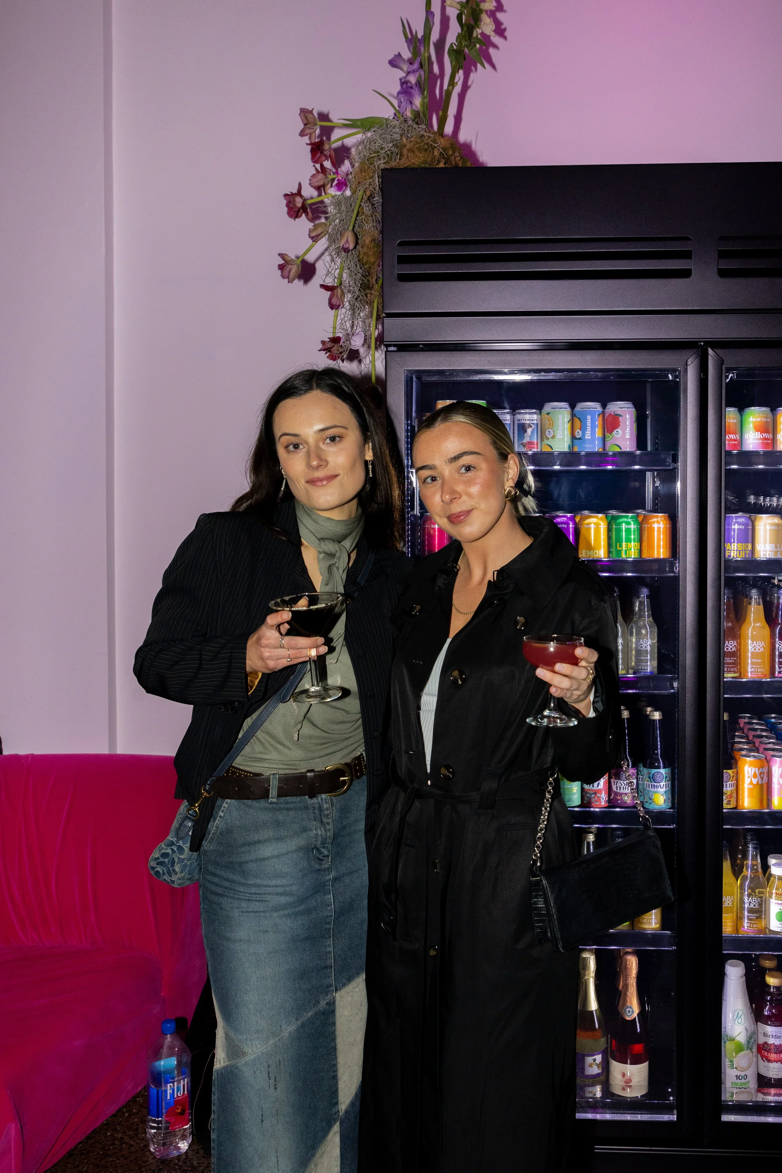 Two women standing in front of a beverage fridge, holding cocktails, with a pink sofa and a water bottle on the floor nearby.