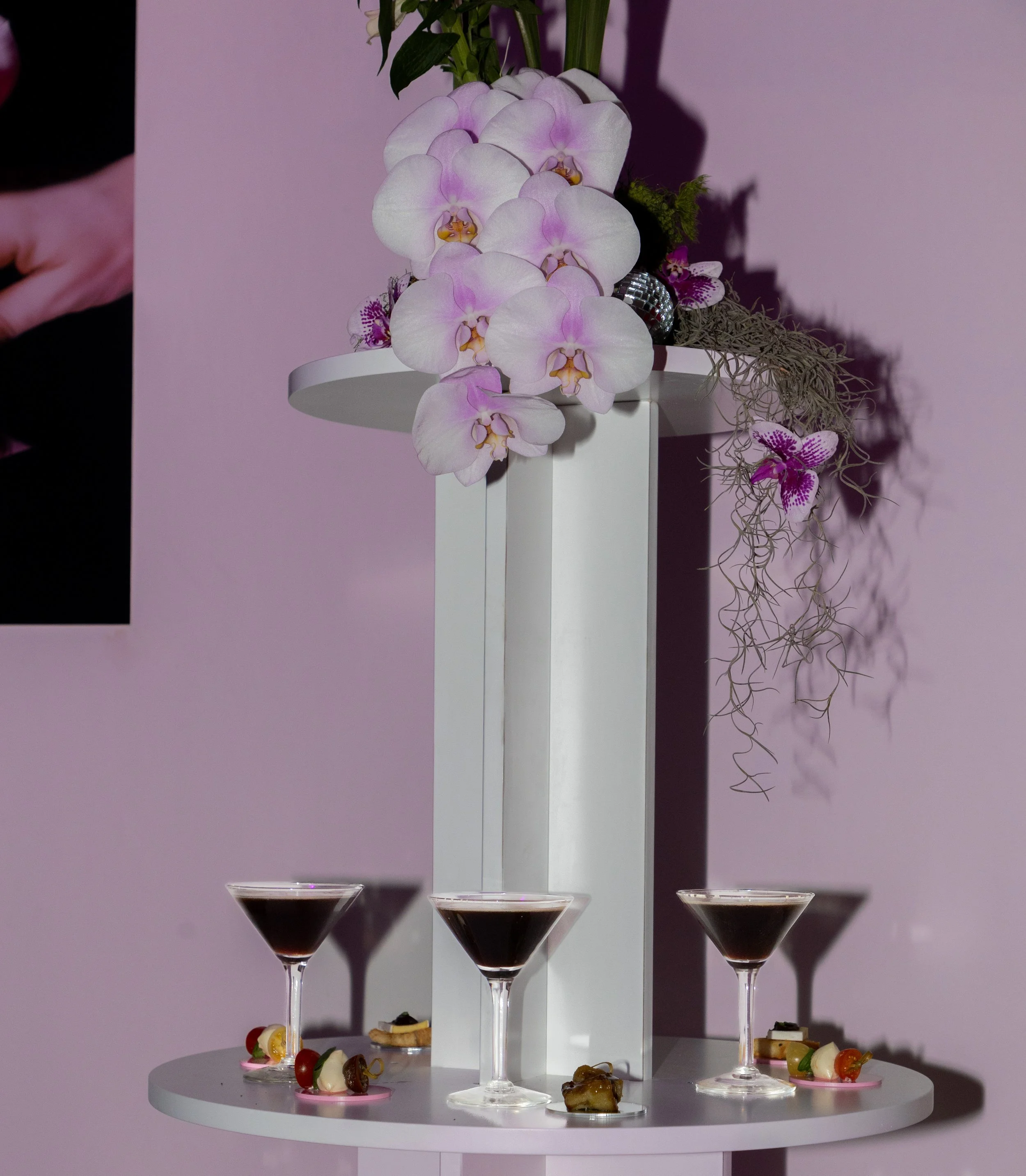 A white table holding three glasses of red wine or cocktail with small hors d'oeuvres, topped with an arrangement of pink and white orchids, purple flowers, and greenery against a light pink wall.