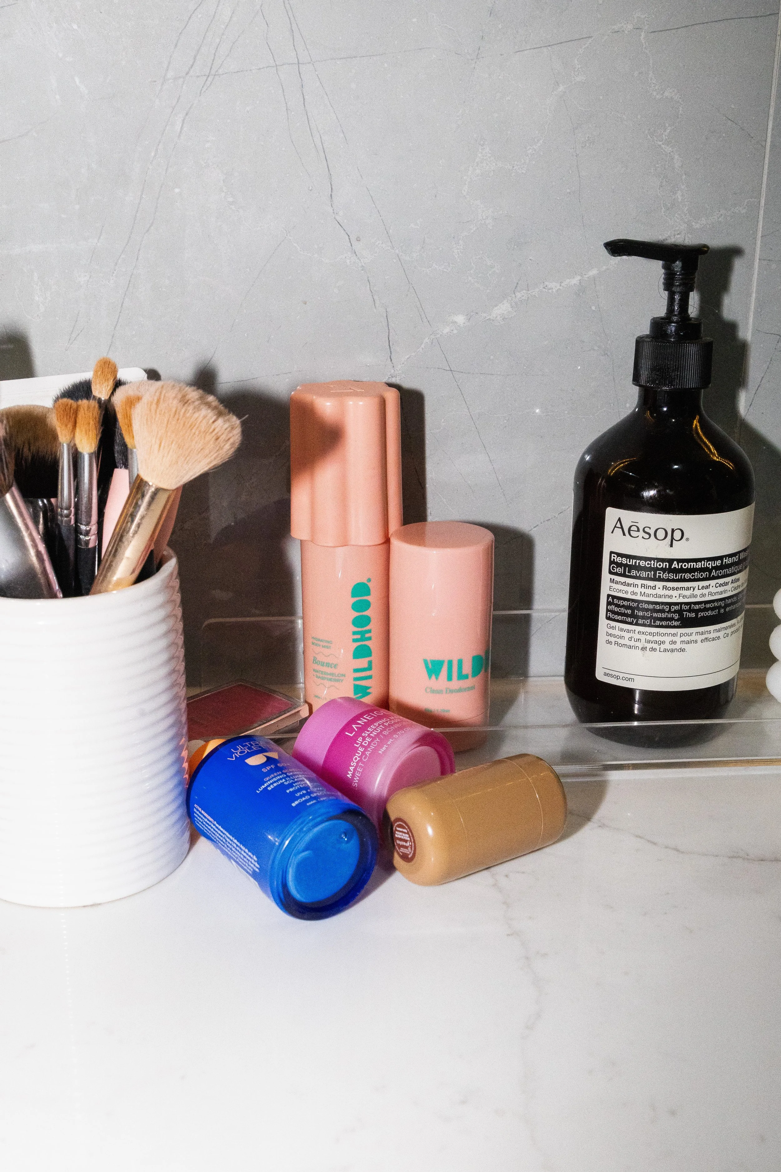 A collection of cosmetic and skincare products including brushes in a white container, pink bottles labeled 'Wild' and 'Wild', blue and pink containers, and a black bottle of Aesop liquid hand soap.