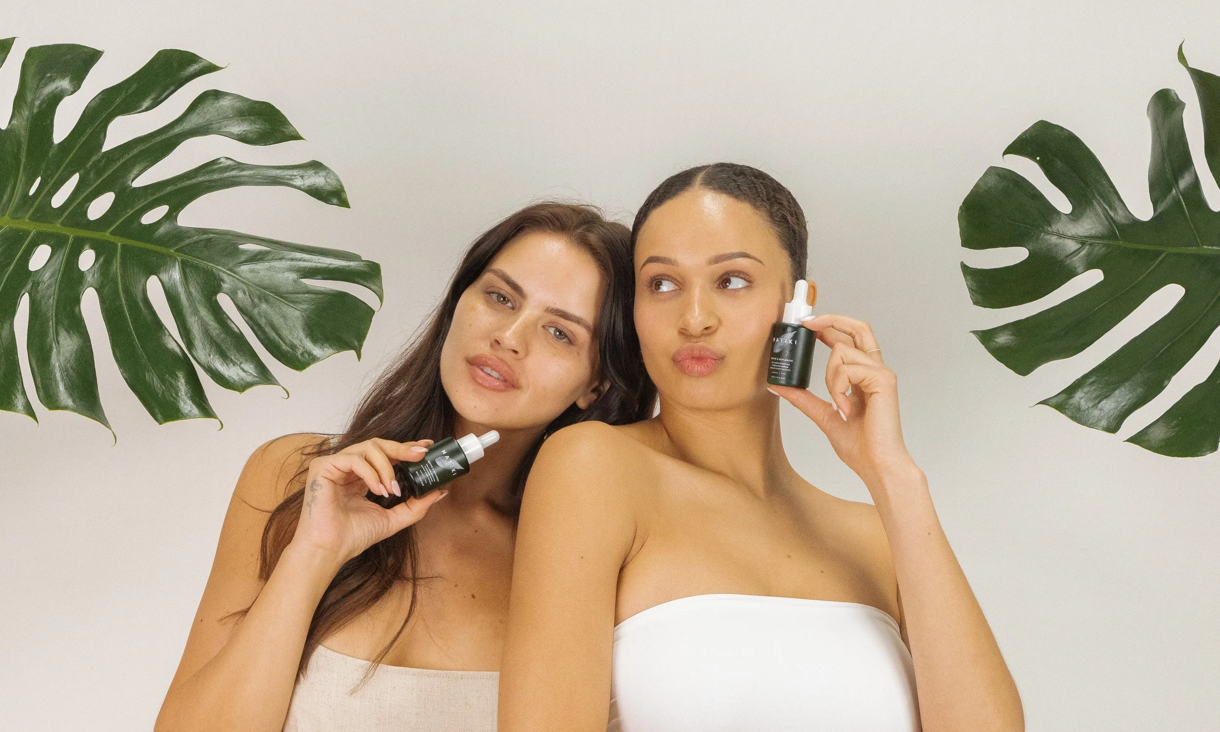 Two women holding skincare serum bottles, with large green tropical leaves on either side, in front of a plain background.