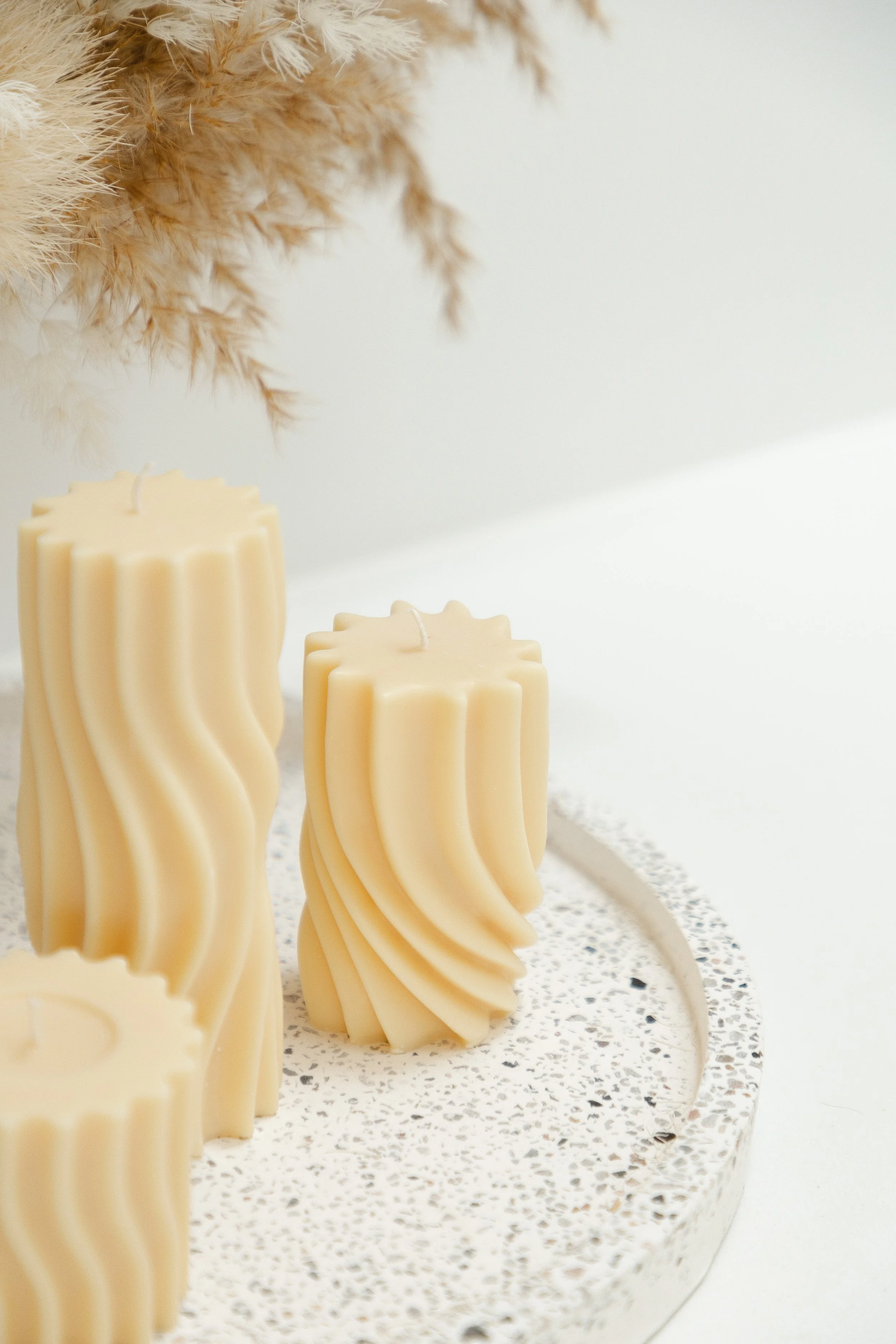Four white pillar candles with wavy design on a speckled white surface, with a dried pampas grass arrangement in the background.