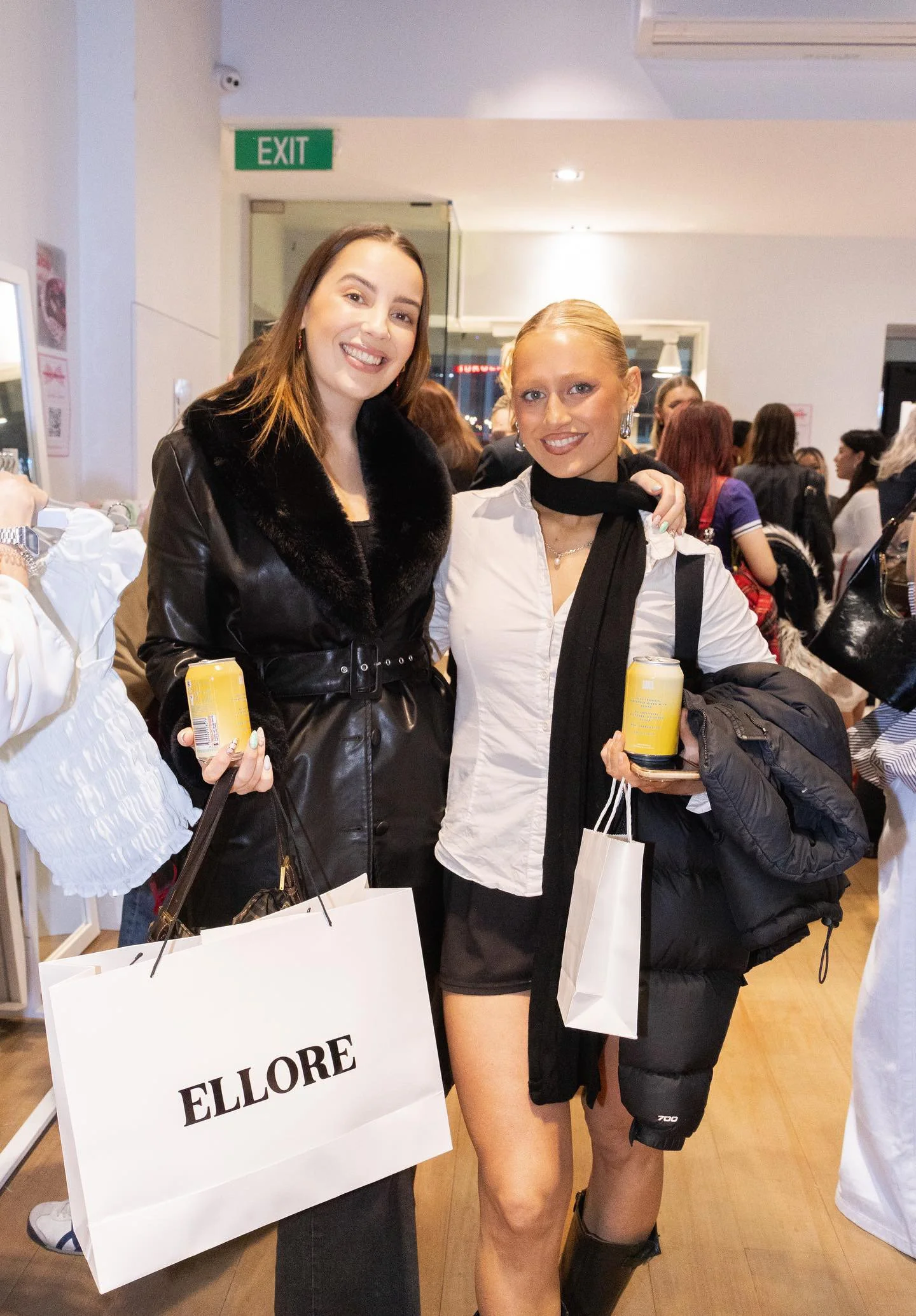 Two women smiling and posing together at a shopping event. One is holding a large shopping bag labeled ELLMORE, and both are holding cans of drinks.