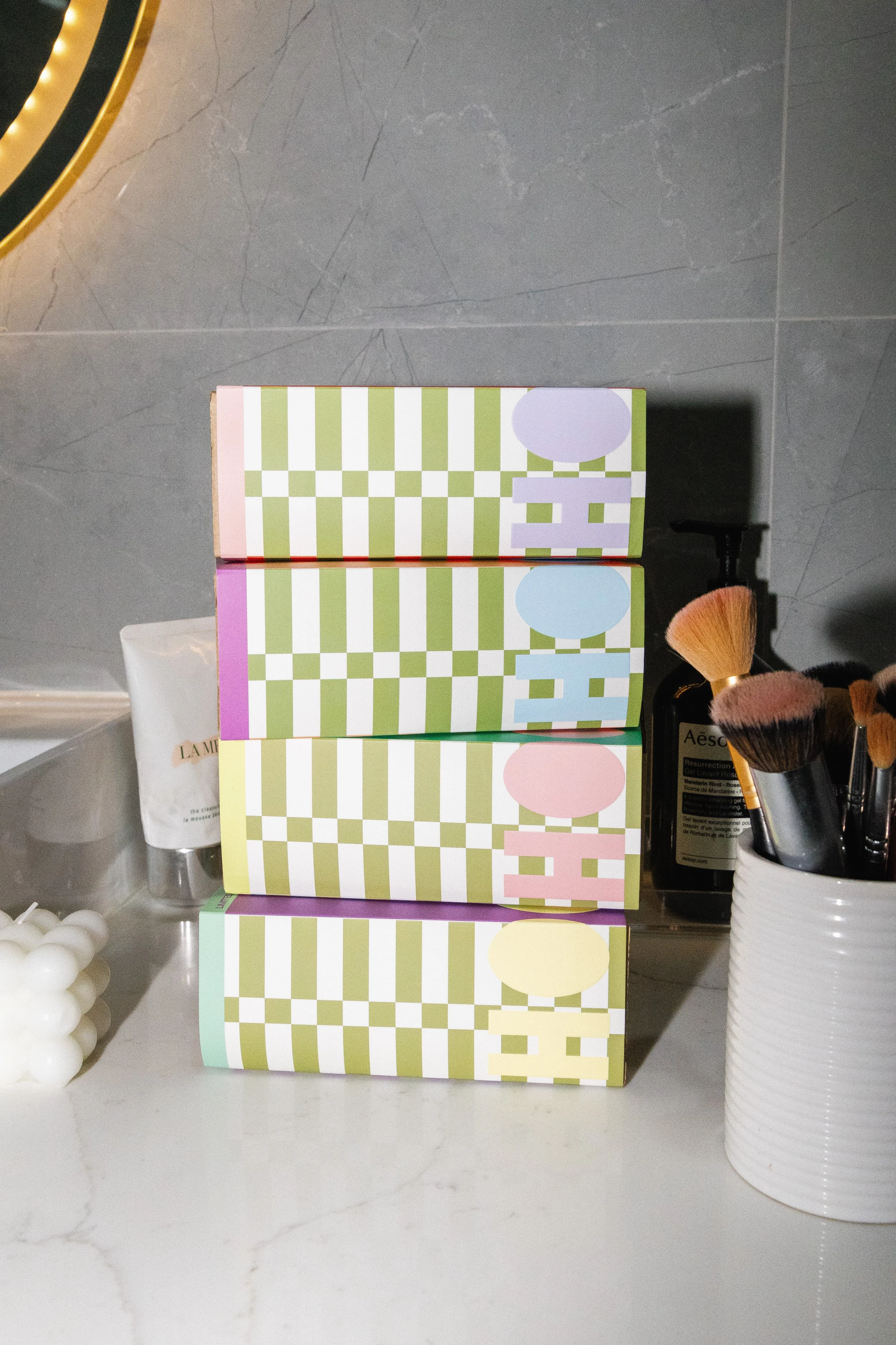 Colorful boxes with checkered patterns in pastel colors stacked on a marble countertop, with makeup brushes in a white ribbed container and a tube of skincare product in the background.
