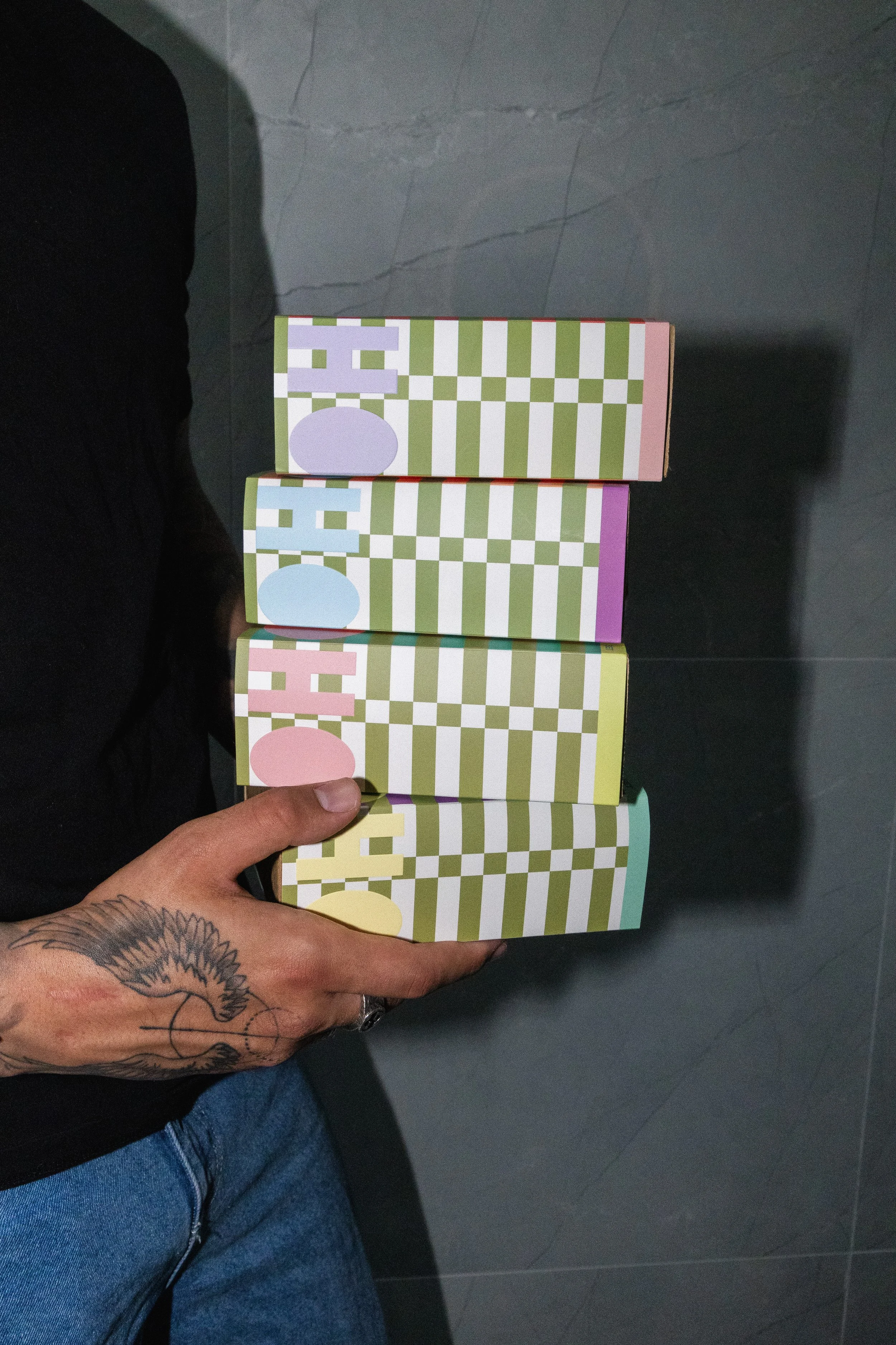 A person with a tattoo on their hand is holding four colorful boxes with checkered patterns and pastel-colored eggs on the sides. The person is wearing blue jeans and a black shirt, standing on a gray tiled floor.