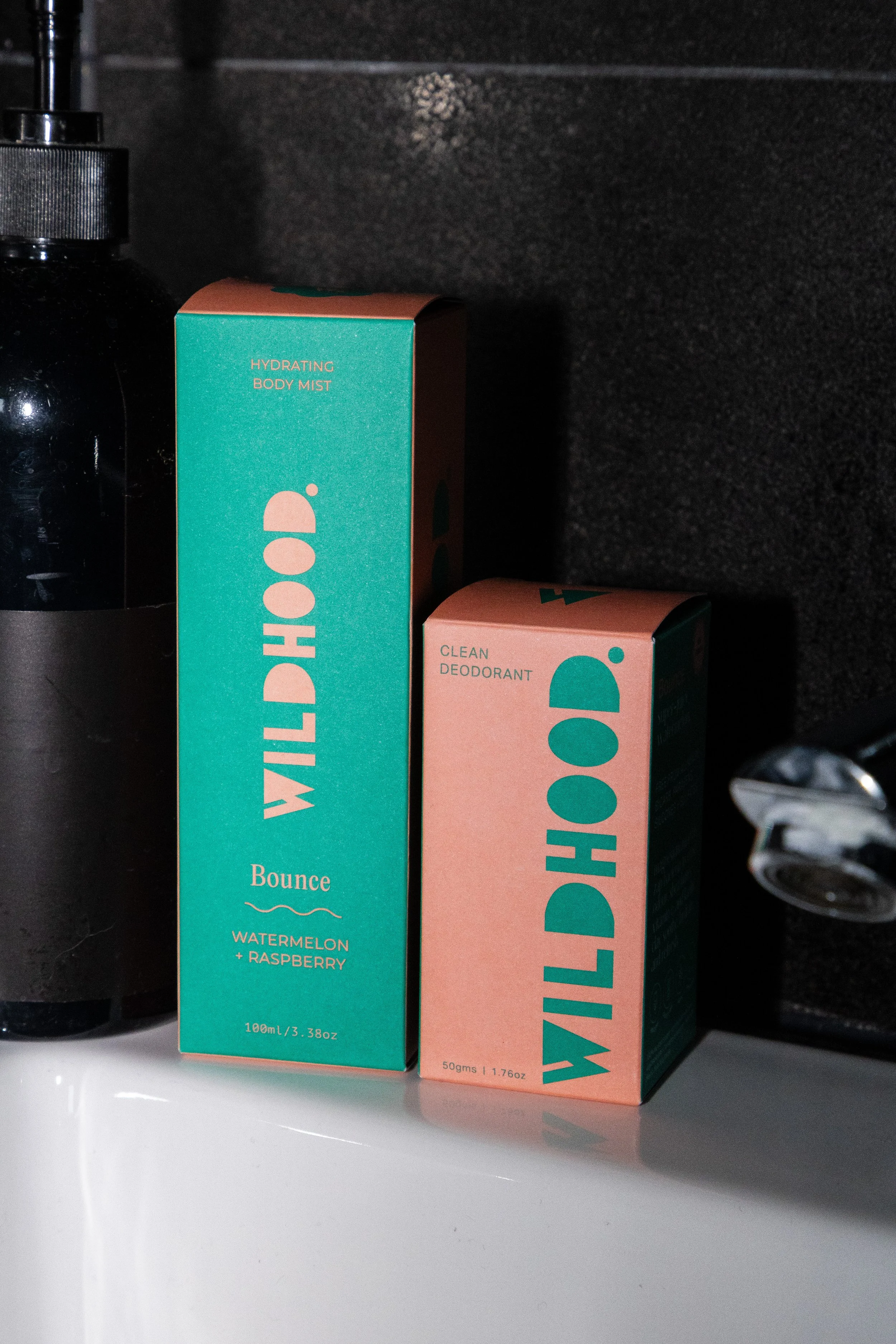 Two Wildhood personal care products, a teal box labeled 'Hydrating Body Mist' with scent 'Watermelon + Raspberry' and a smaller pink box labeled 'Clean Deodorant,' positioned on a white surface next to a black bottle and a metallic faucet handle, aga