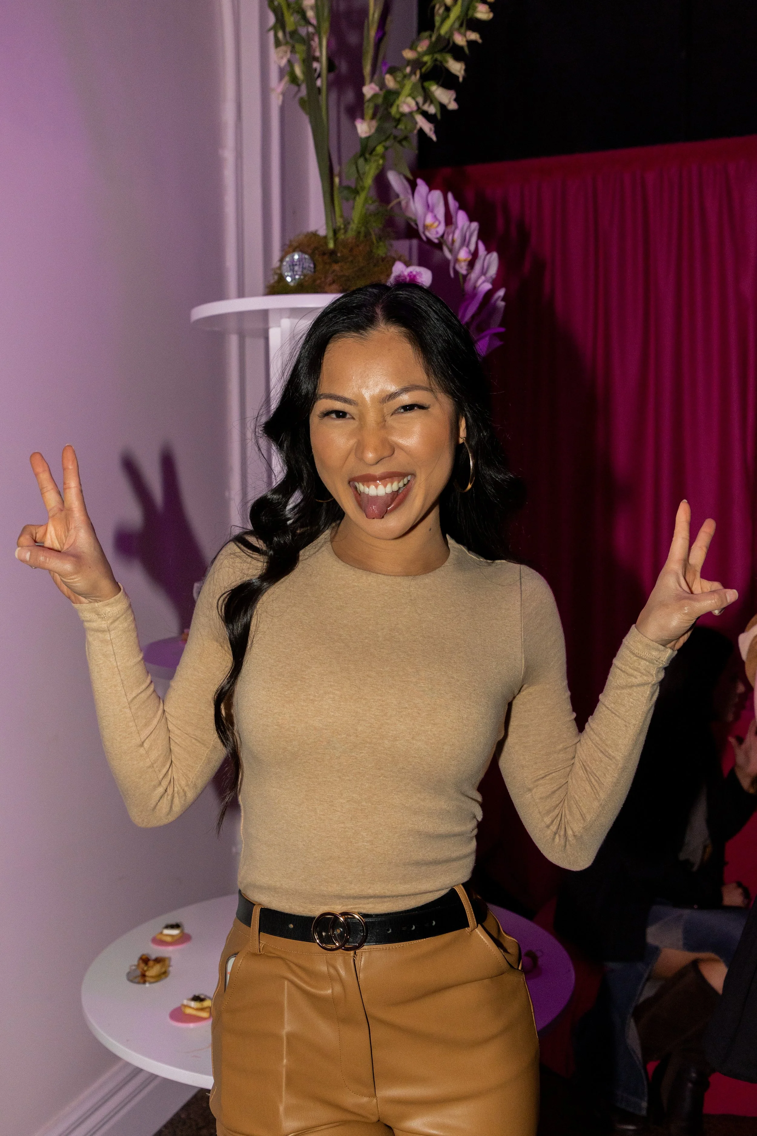 A woman with long black hair doing peace signs with both hands, sticking her tongue out, and smiling at the camera. She is wearing a beige long-sleeved top, tan pants, and a black belt. There are purple and white flowers in a vase and some snacks on