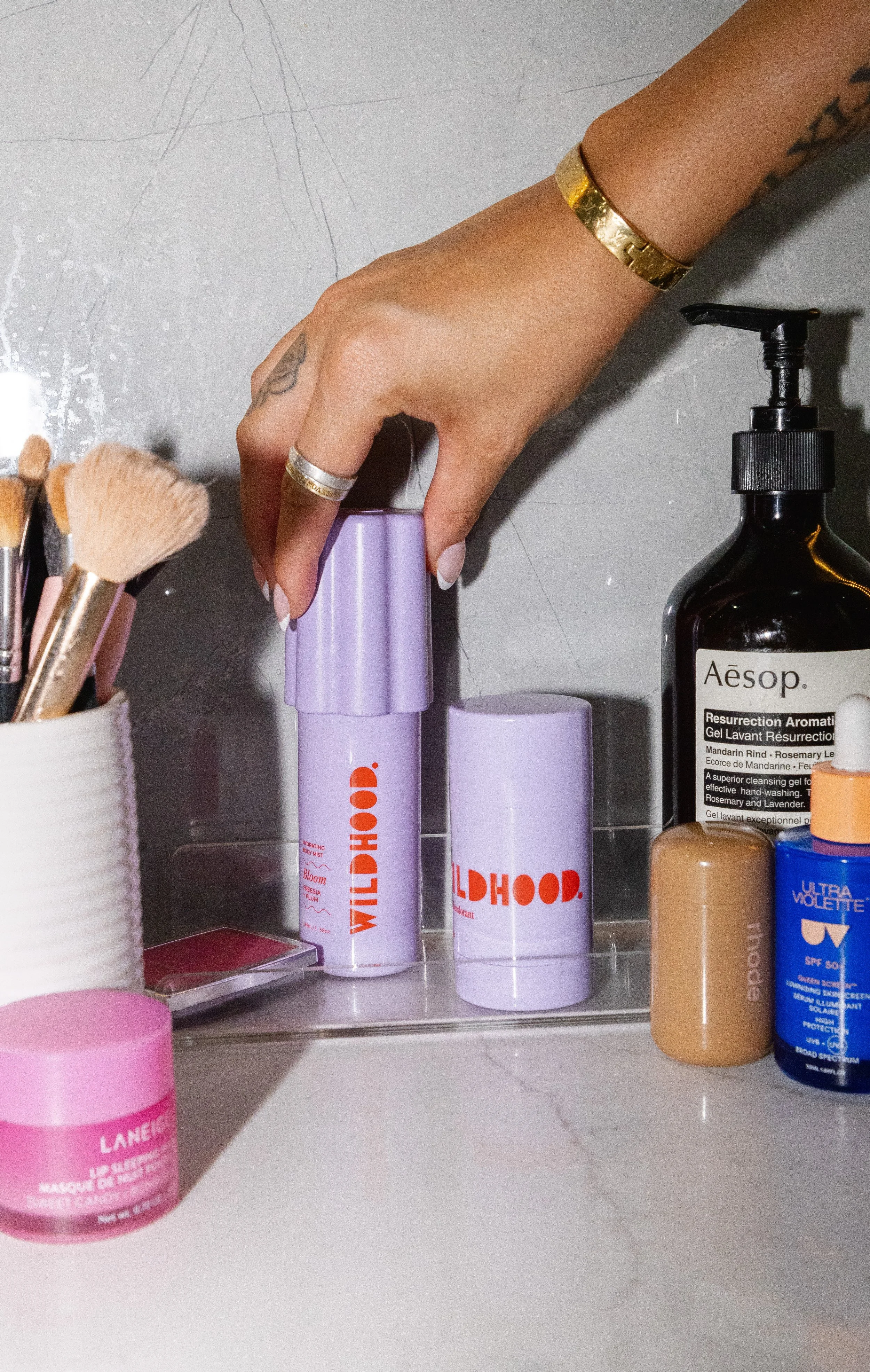A hand with rings and a bracelet reaching for a purple Wildhood beauty product on a bathroom countertop, surrounded by skincare and cosmetic items including brush holders, a large black Aesop bottle, and a pink Laneige jar.