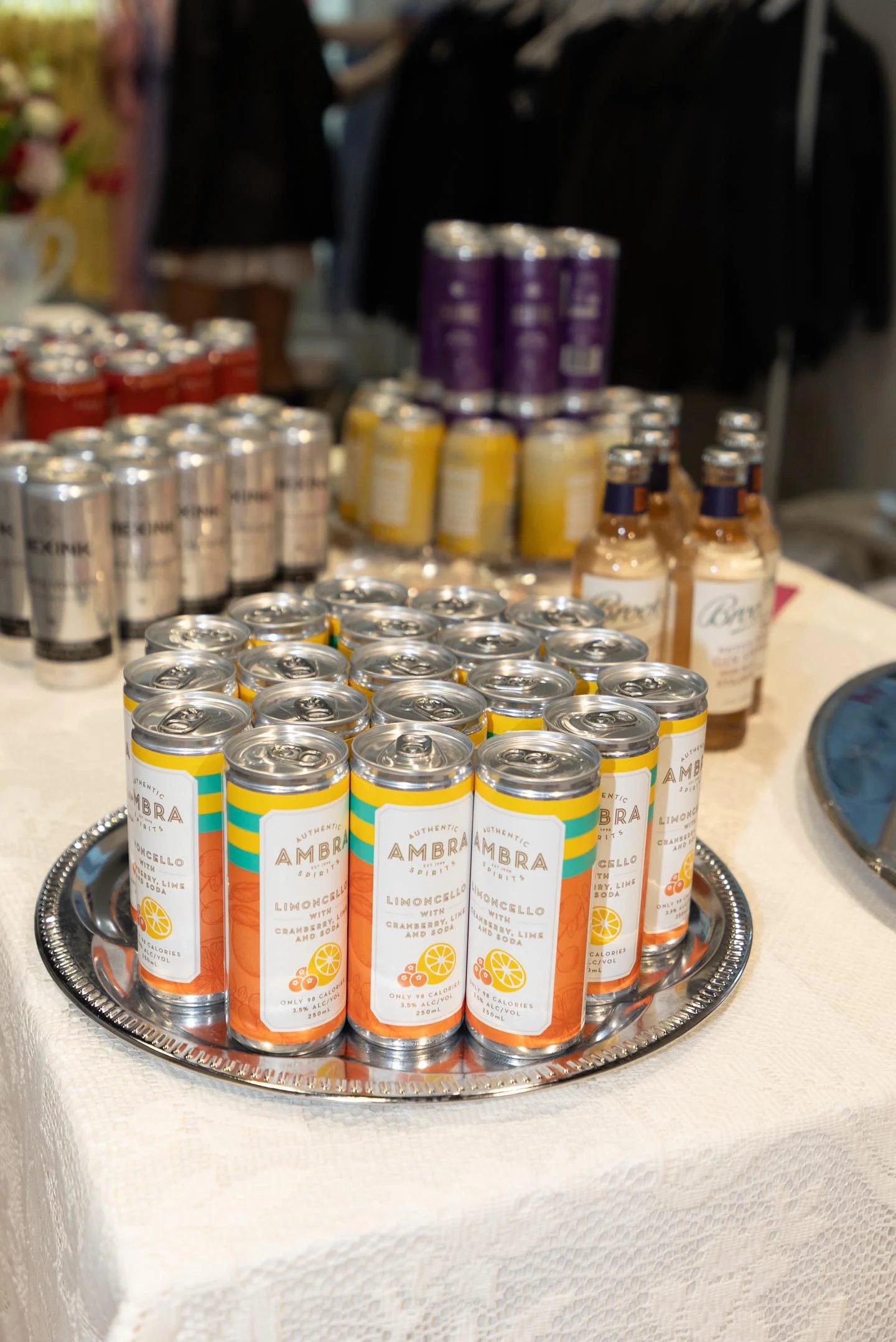 A tray of canned Ambra Limoncello soda with citrus illustrations in the foreground, with other cans and bottled beverages in the background on a table.