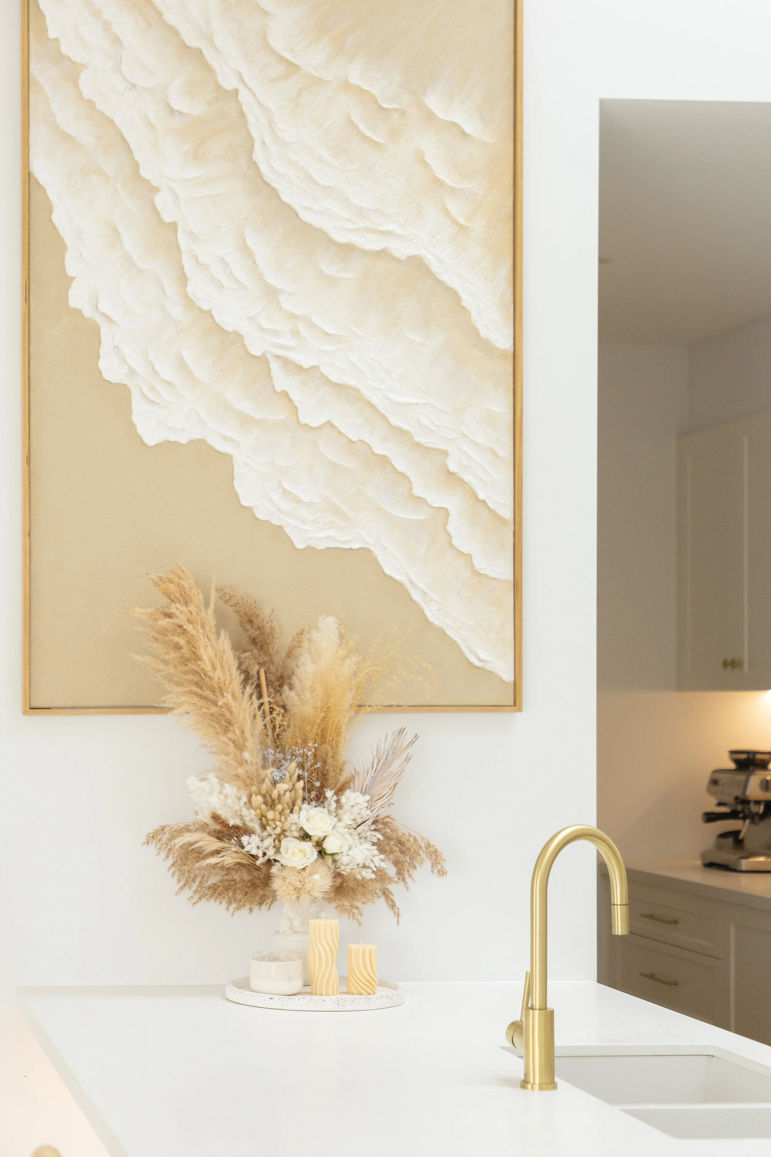 Decorative kitchen wall art with a 3D cloud design, a vase with dried pampas grass and white roses, and a gold kitchen faucet.
