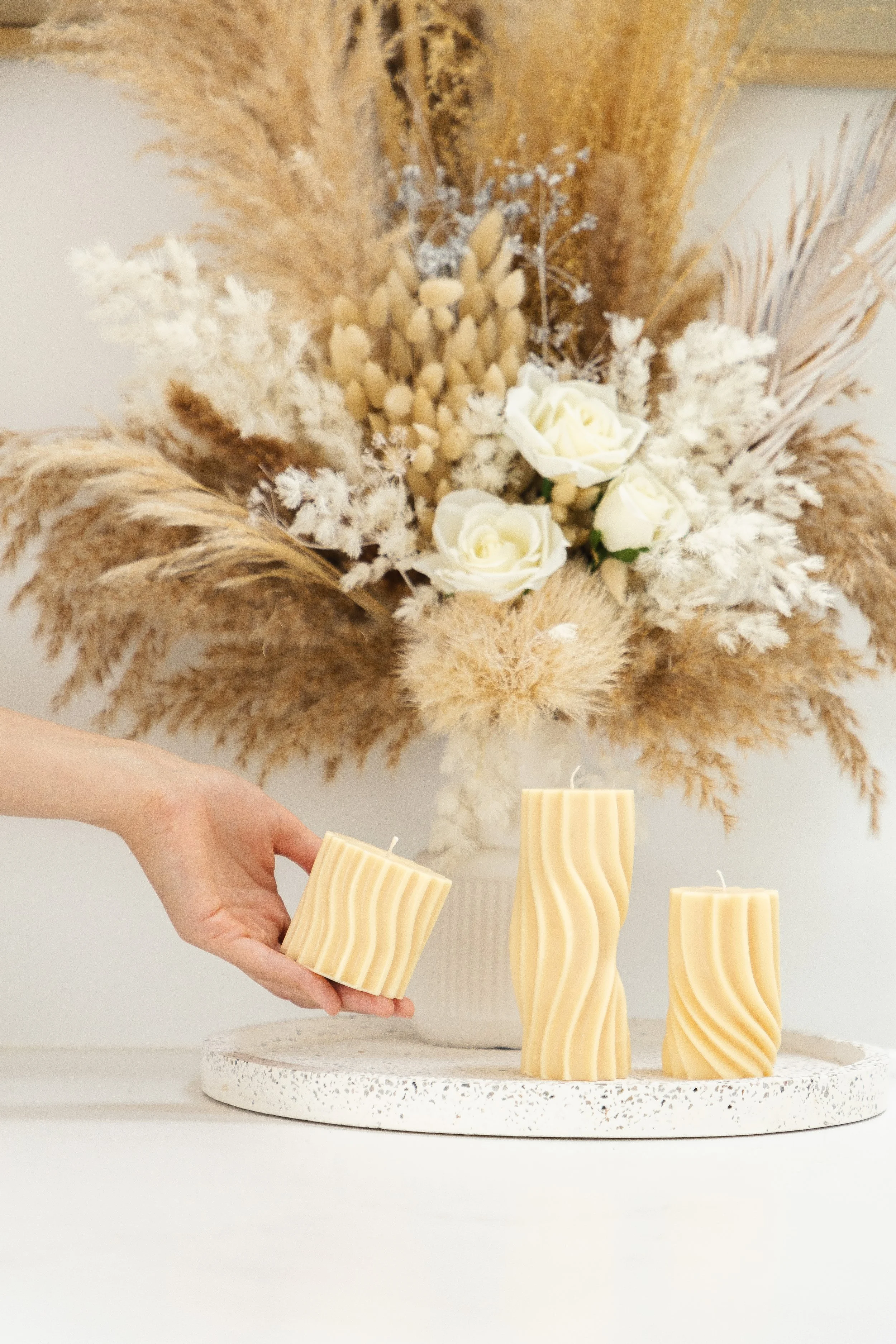 A hand placing a striped cream-colored candle on a white textured tray, with two additional similar candles on the tray, in front of a large neutral-colored floral arrangement with white roses and dried grasses.