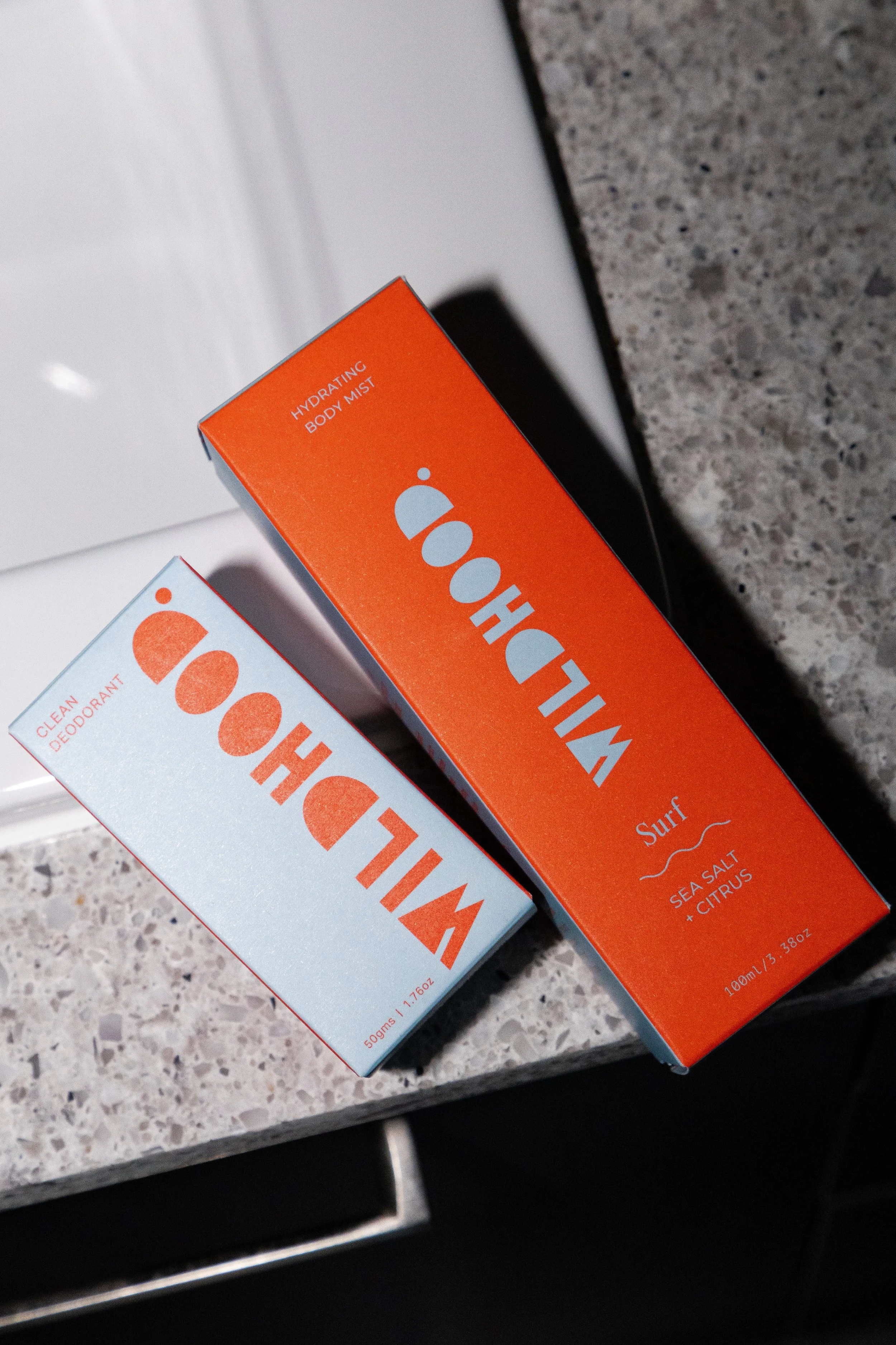 Two boxes of Oooh! brand skincare products on a speckled countertop. One is labeled Hydrating Body Mist in orange packaging, and the other is labeled Clean Deodorant in white packaging.