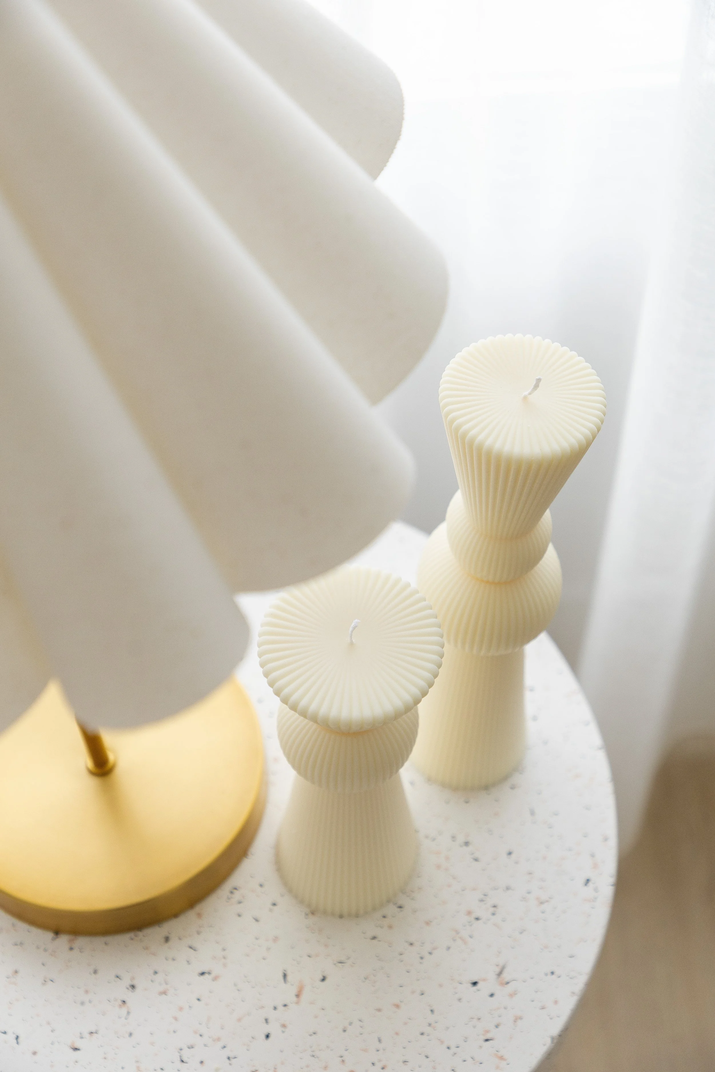 Decorative white candles with textured, ribbed surfaces on a white speckled table near a white curtain, illuminated by soft natural light.