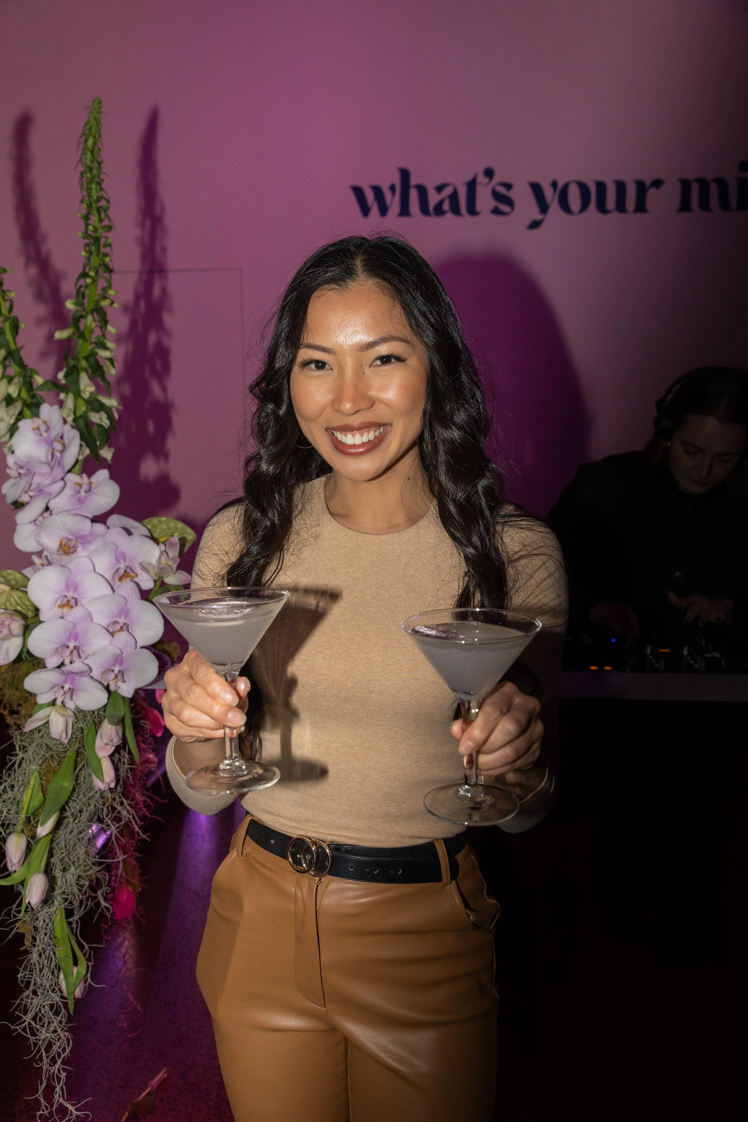 A woman smiling and holding two cocktails in a dimly lit venue with purple lighting. There are flowers on her left and a DJ in the background. The partially visible text on the wall says "what's your mi".