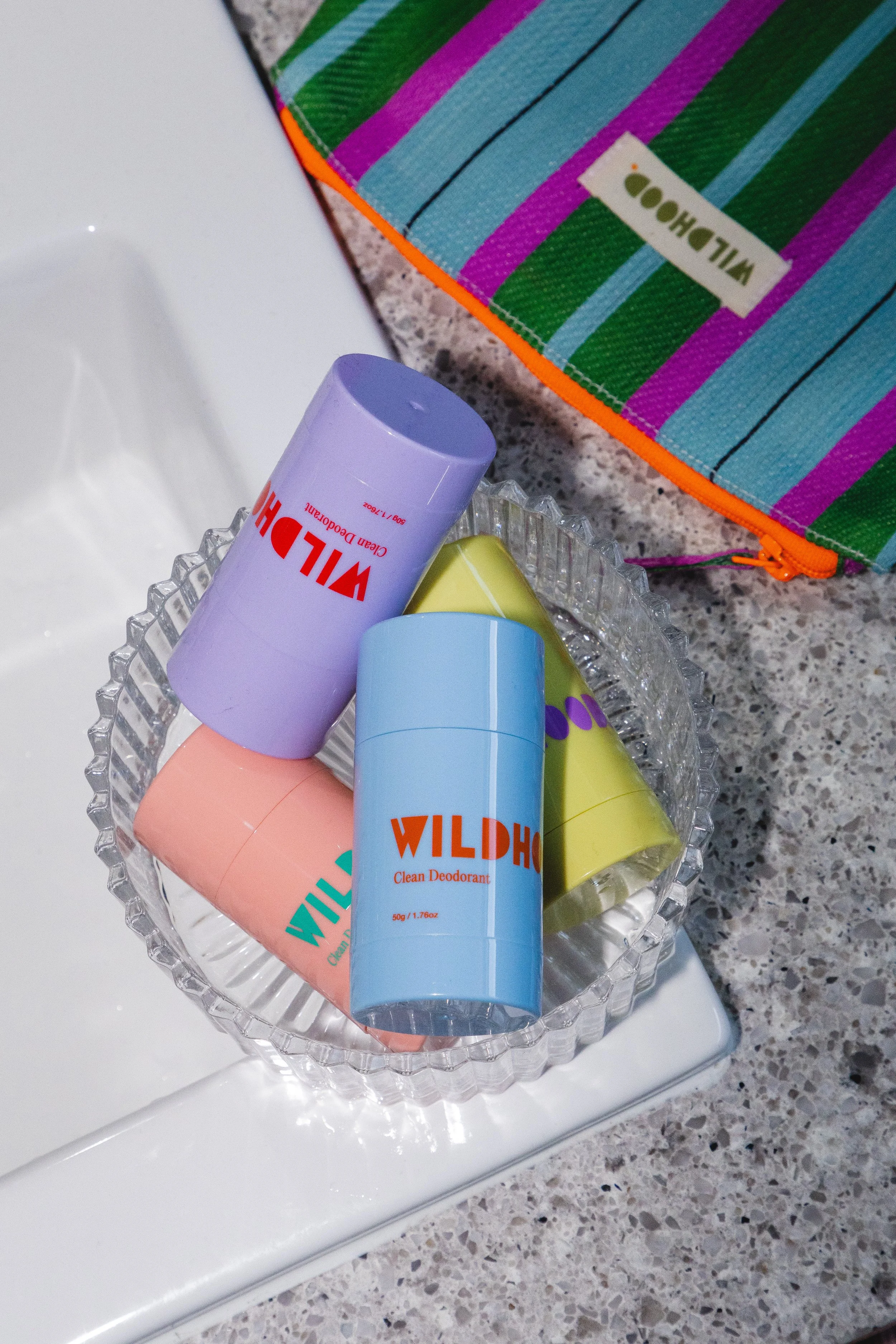 Colored deodorant sticks in a glass dish on a counter with a colorful striped bag nearby.