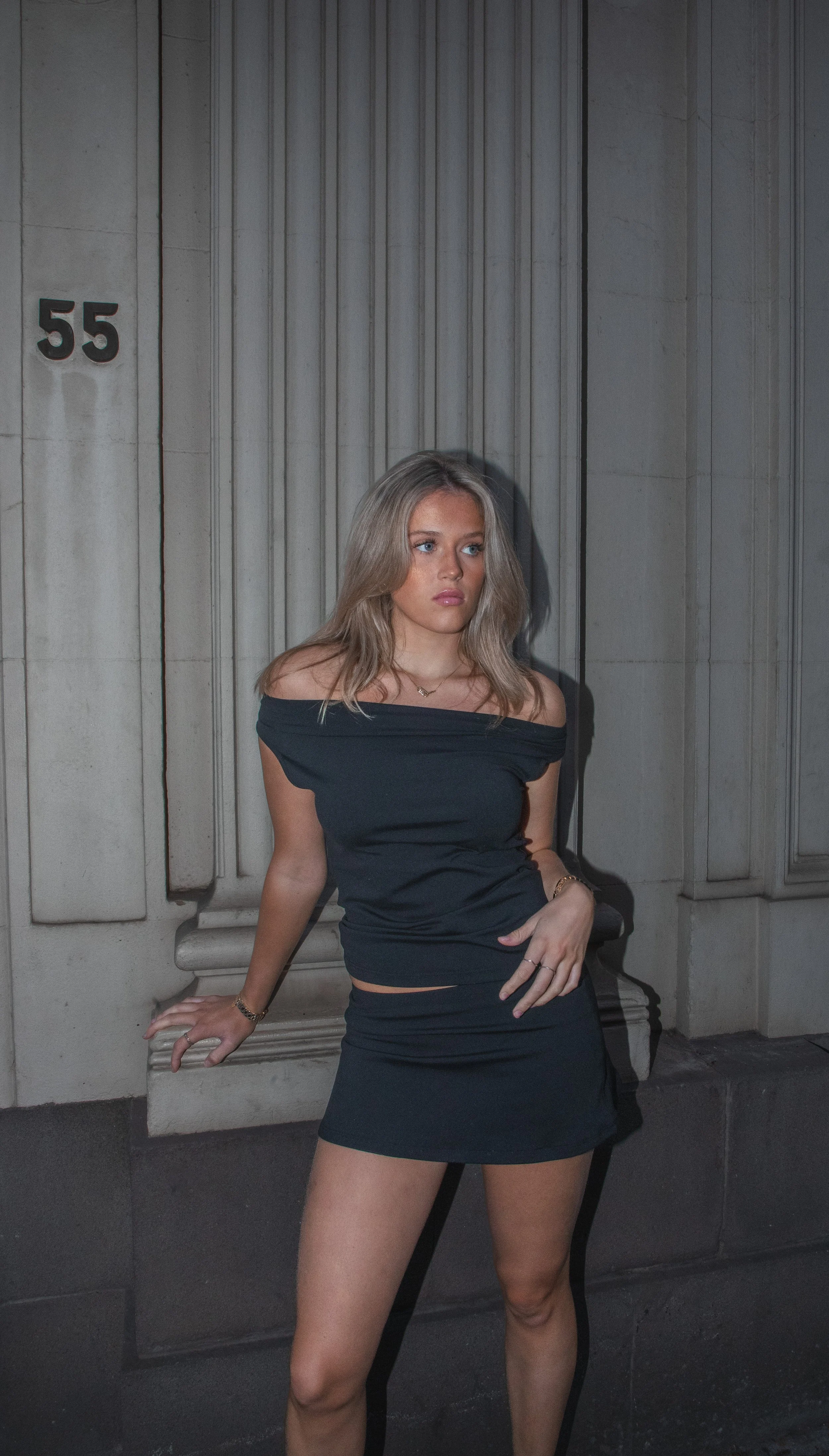 A woman with long blonde hair wearing a black off-the-shoulder dress standing against a stone wall with the number 55 on it.