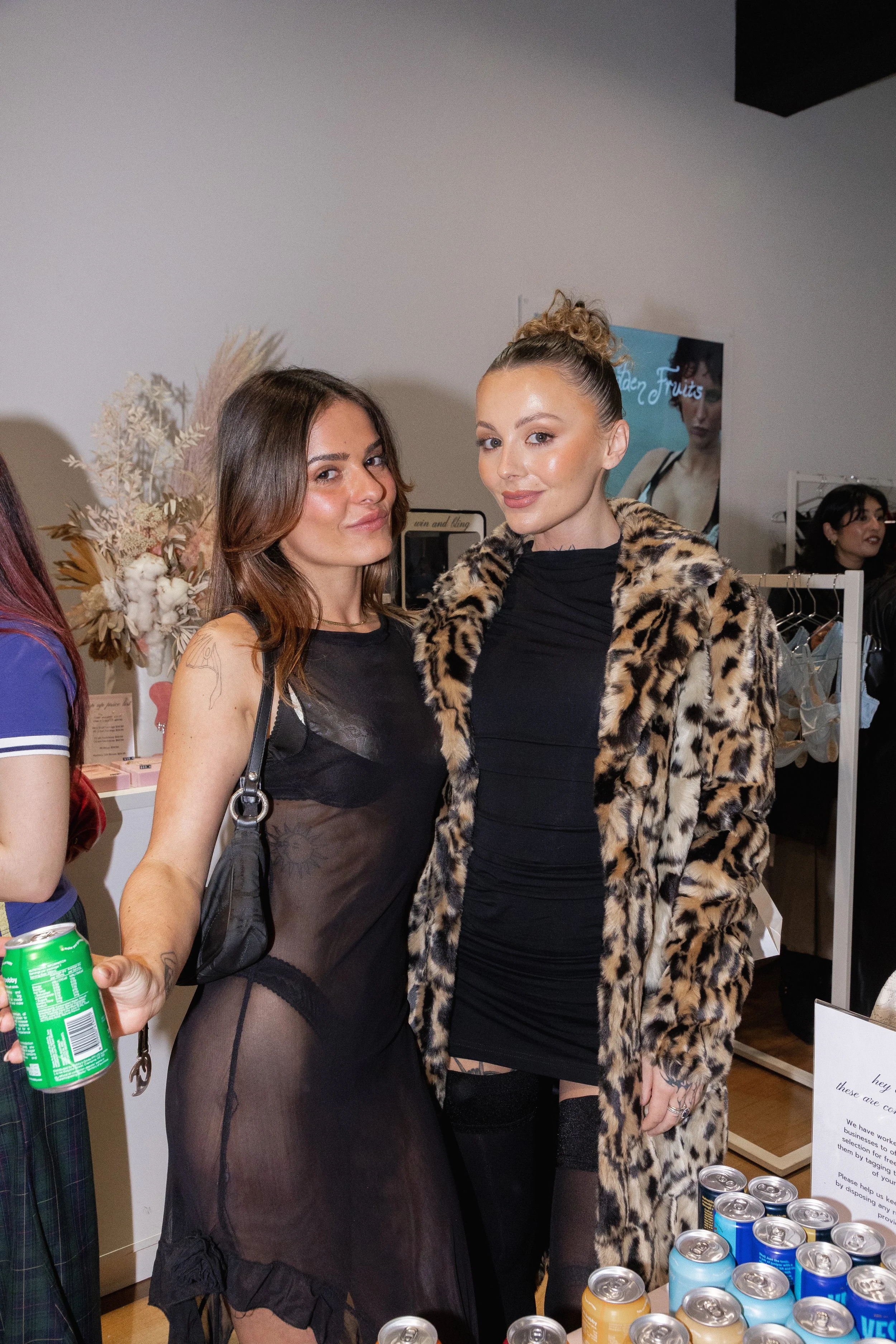 Two women standing together at a social event, one wearing a sheer black dress and the other wearing a black dress with a leopard print coat.