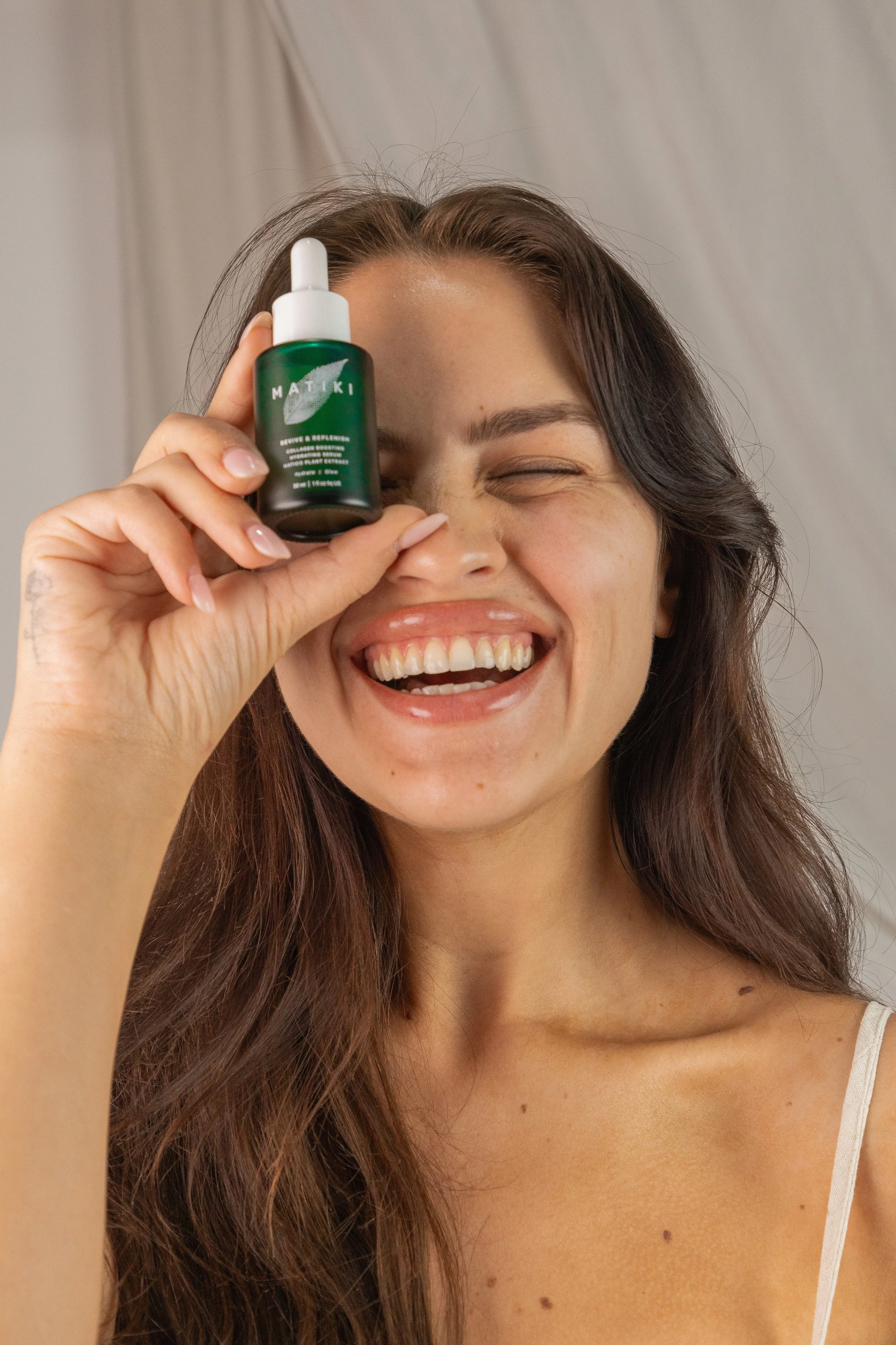 Smiling woman with long brown hair holding a small green bottle of skincare serum up to her eye, standing in front of a neutral background.