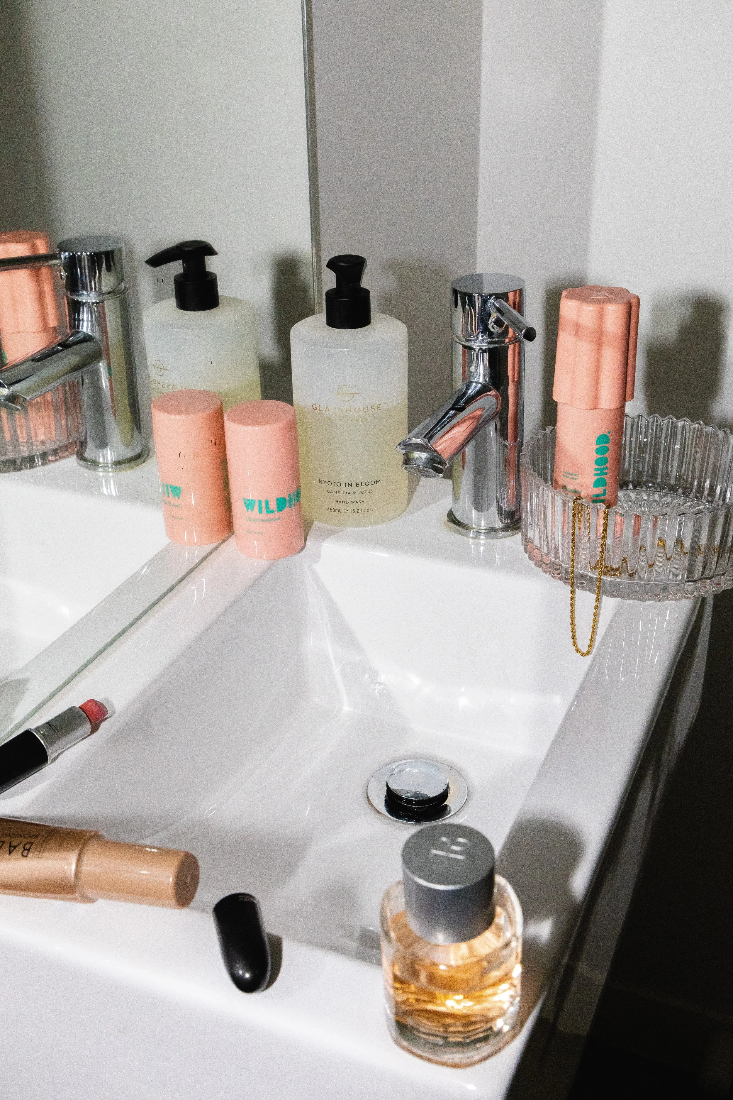 Bathroom sink with various skincare and beauty products, including soap dispensers, perfume, lipstick, and a mirror.