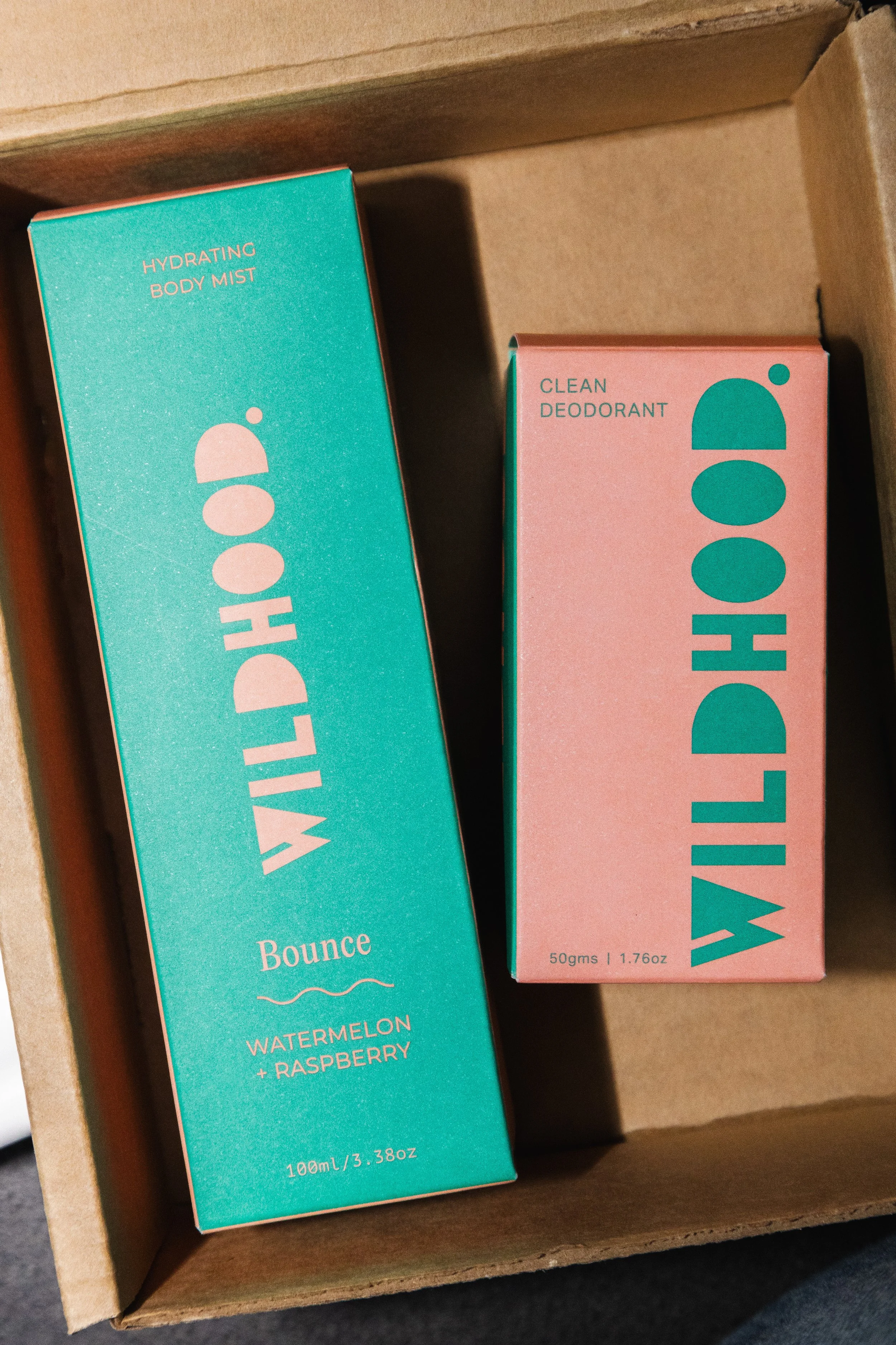 Two boxes of Willdhoo health and wellness products in a cardboard box. The teal box on the left is labeled 'Hydrating Body Mist Bounce Watermelon + Raspberry' with a volume of 100ml. The pink box on the right is labeled 'Clean Deodorant' with a weigh