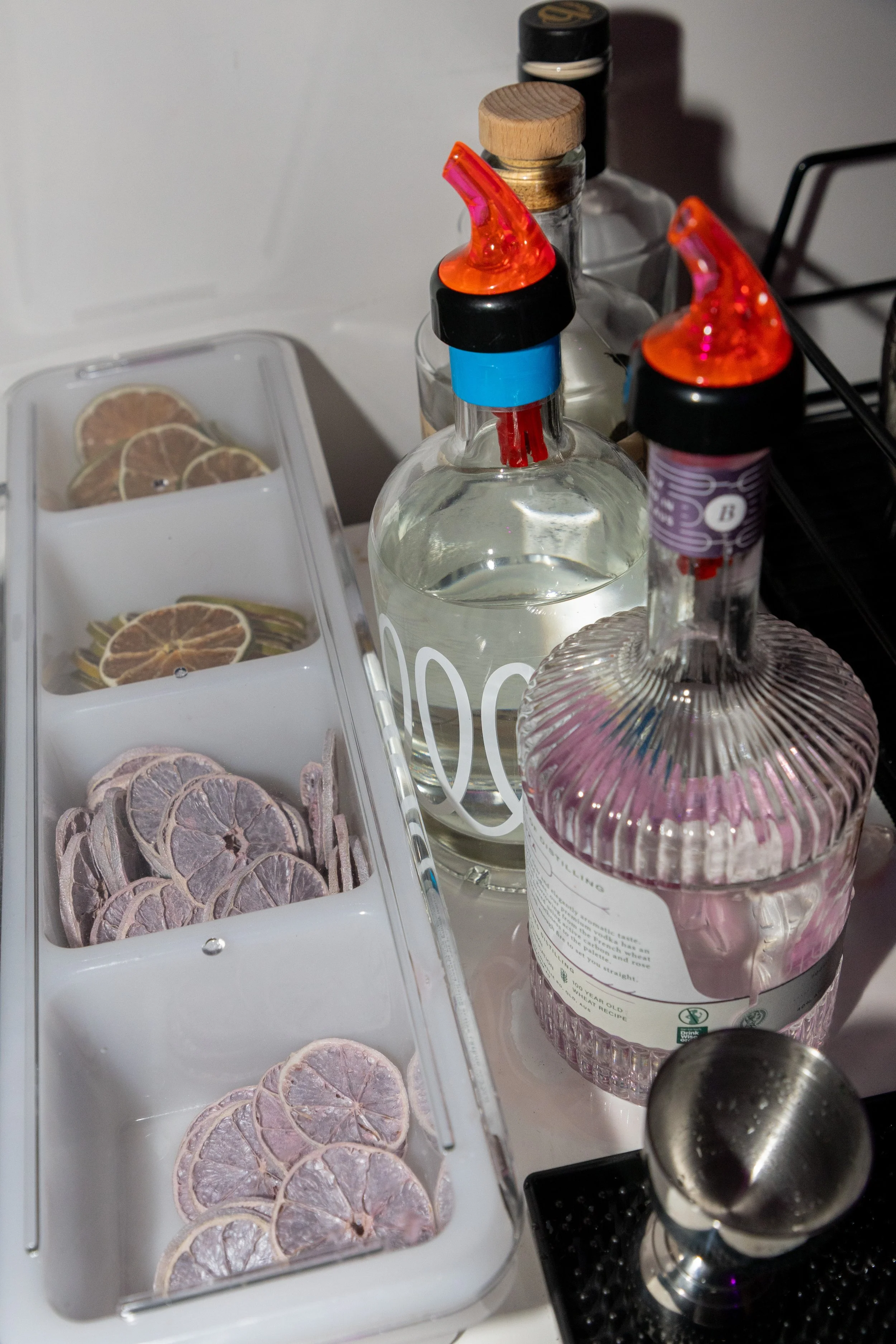 A white ice tray with dried citrus slices, several bottles of alcohol including flavored spirits with red chili pepper infusions, and a metal jigger, all on a kitchen counter.