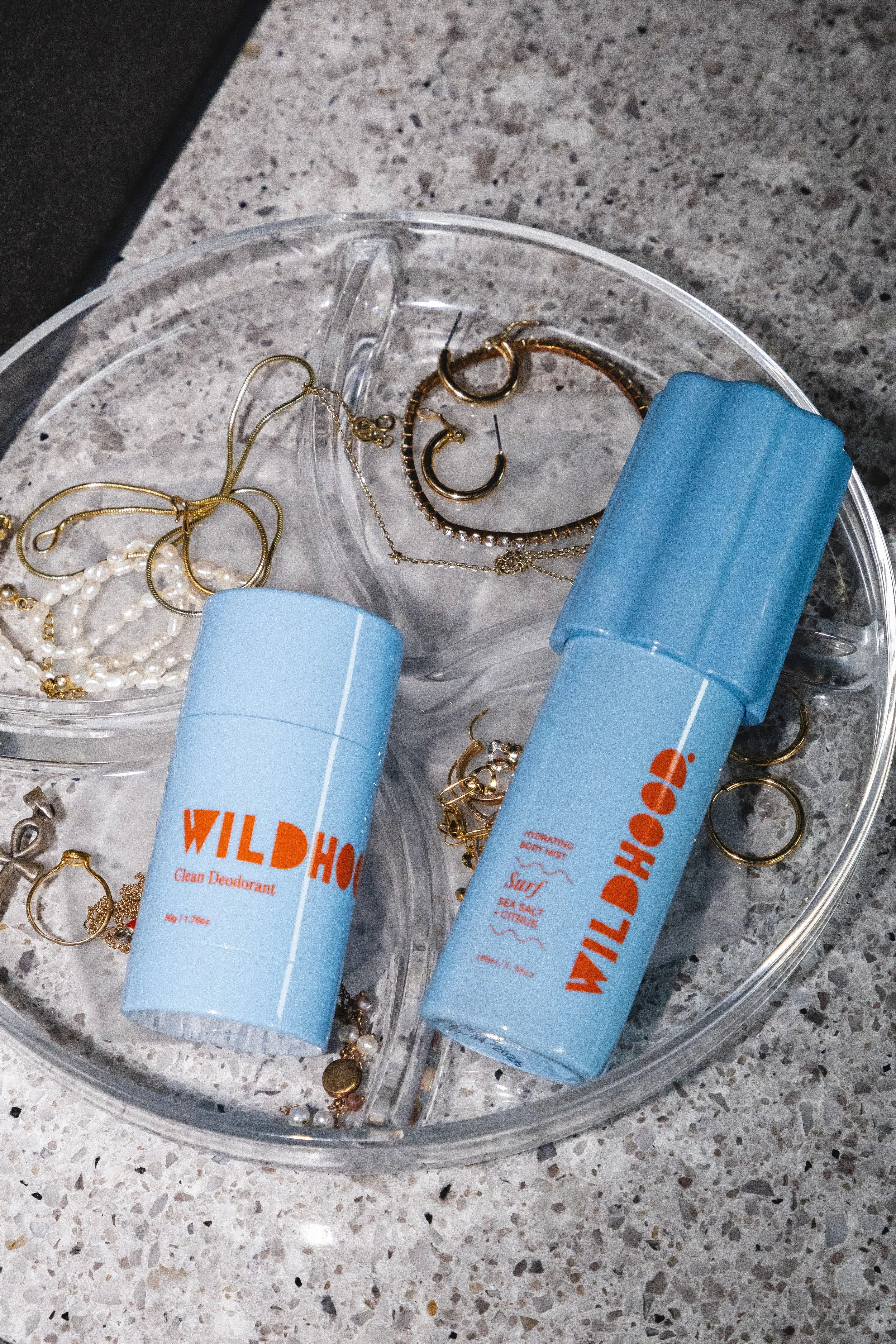 A clear plastic jewelry organizer with compartments holding jewelry and two blue Wild Horn body deodorant sprays on a speckled countertop.