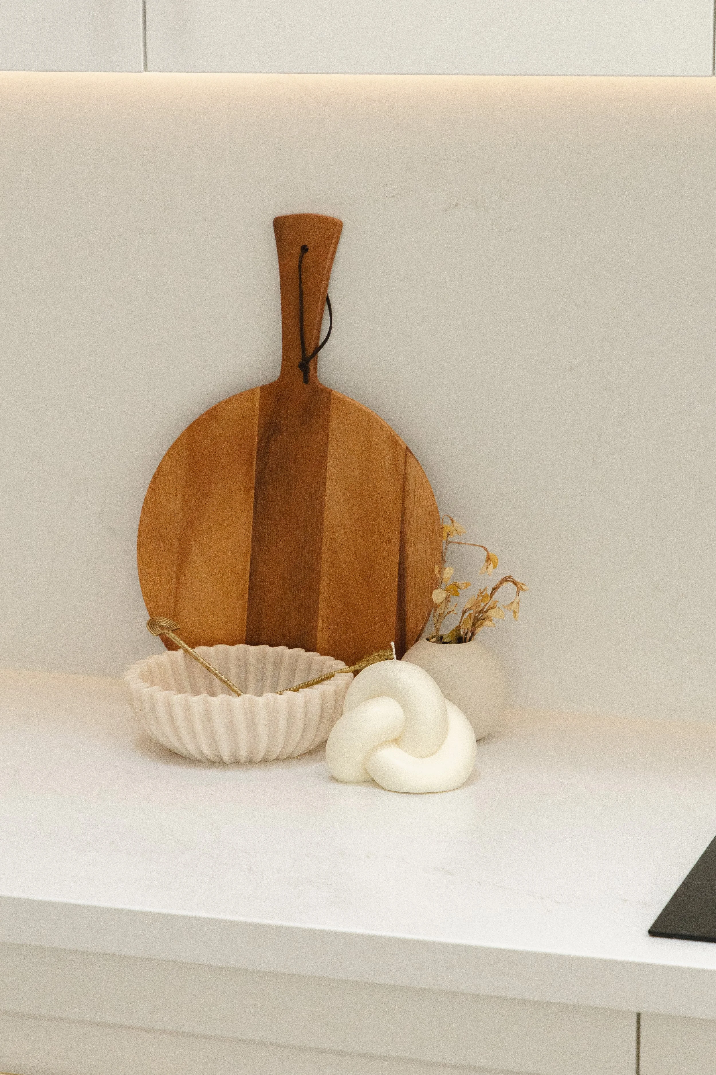 Decorative objects on a white surface, including a round wooden cutting board, a white ceramic bowl with a rope handle, a white knot-shaped sculpture, a small vase with dried flowers, and part of a black item on the right edge.