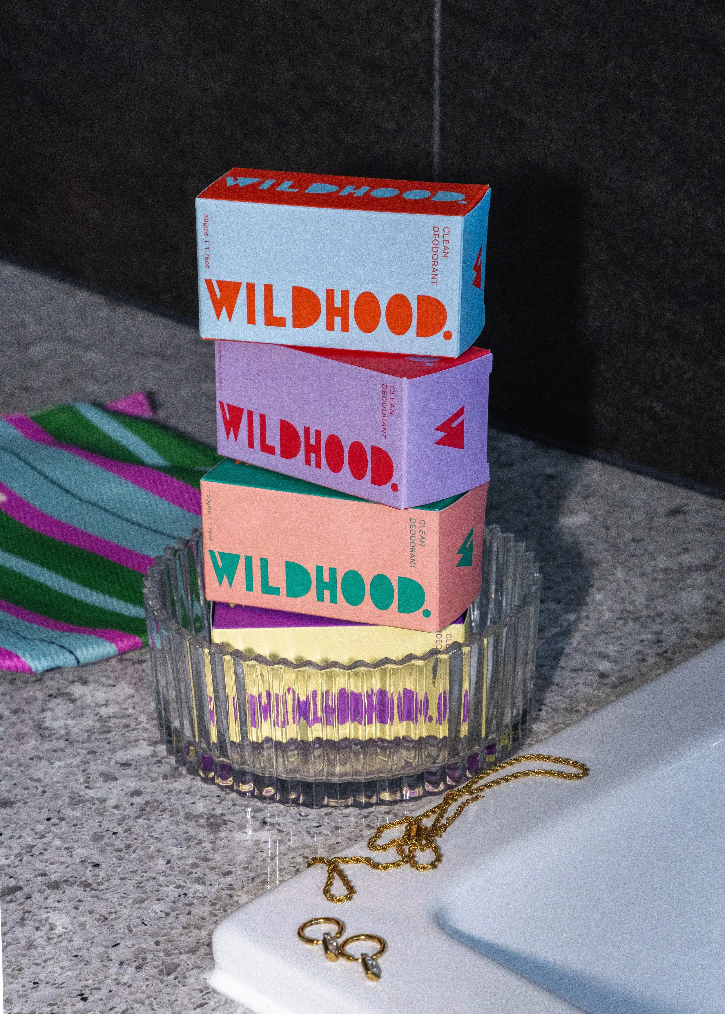 Colorful boxes labeled 'Wildhood' stacked in a glass dish on a kitchen countertop, next to a white sink, a gold chain necklace, and a striped cloth in the background.