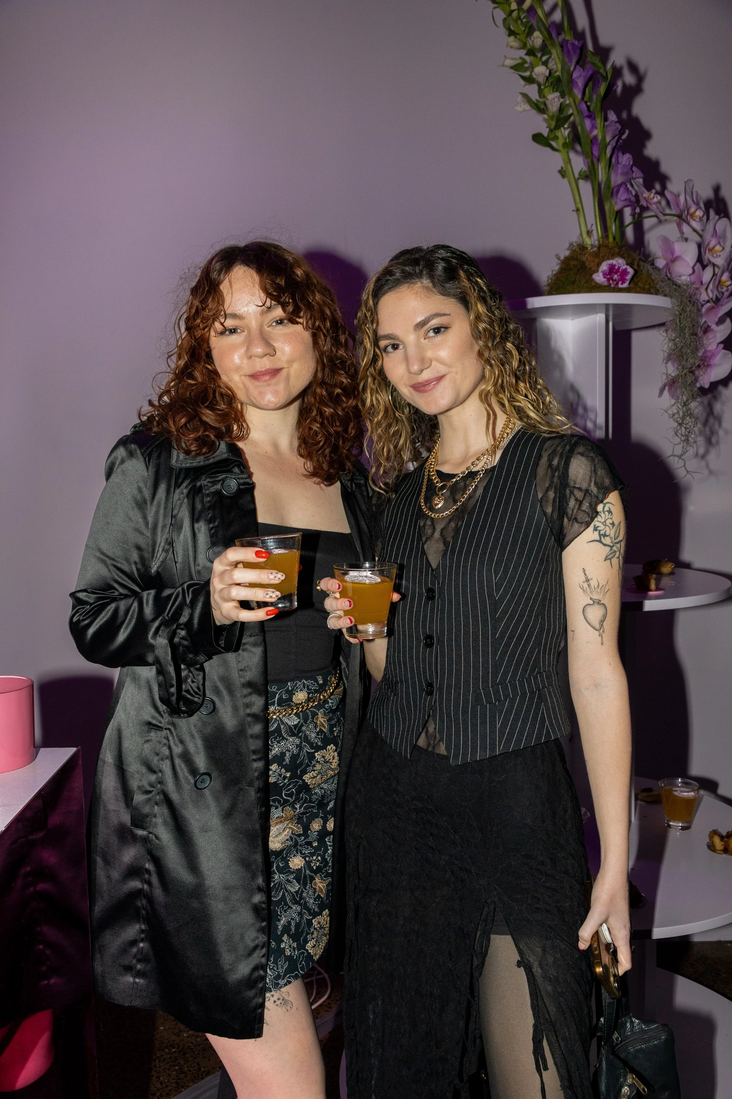 Two women standing close together, holding drinks, smiling at the camera at a social event. They are dressed stylishly, with one in a black satin coat and the other in a black striped vest and skirt. The background features purple lighting and floral