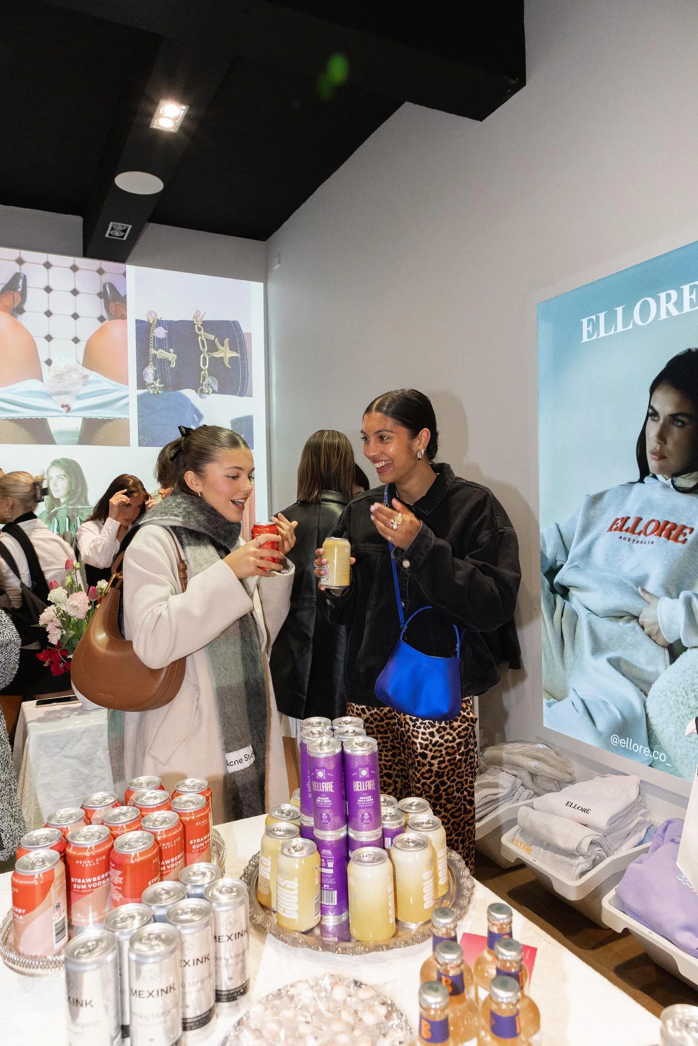 Two women enjoying drinks at a social event with a display of assorted canned beverages on the table, and a large advert featuring a woman in casual clothing on the wall behind them.
