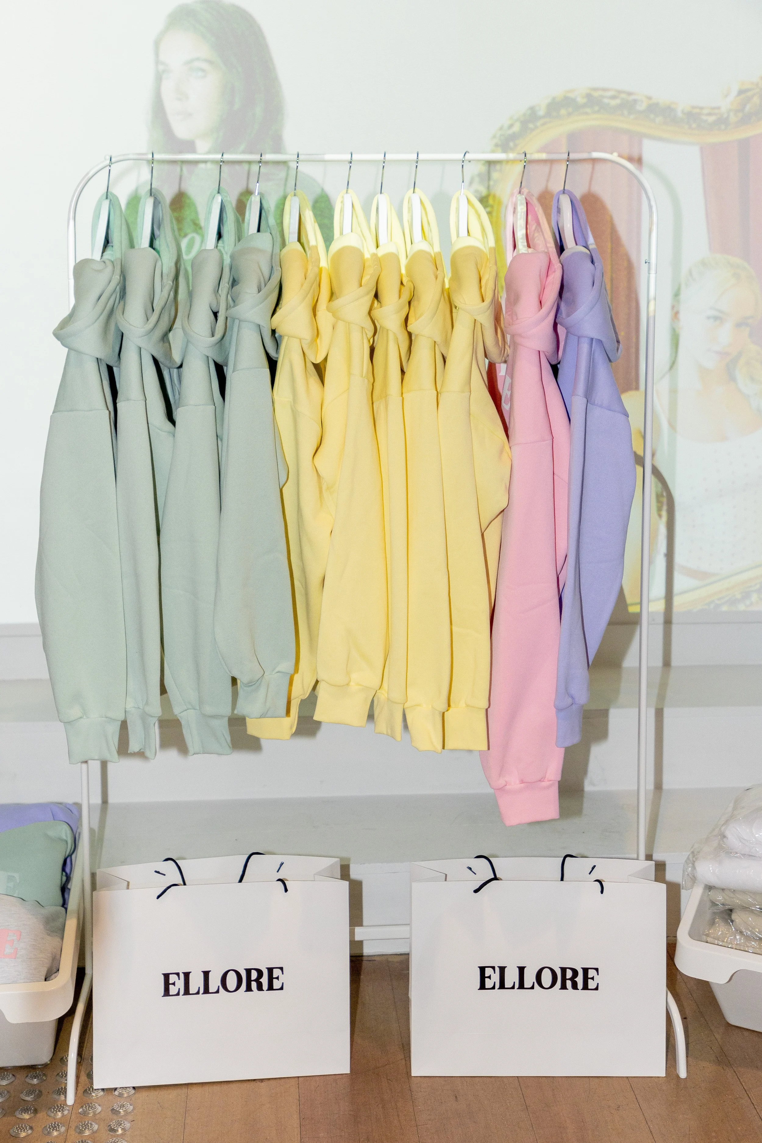 A clothing rack with pastel-colored sweatshirts in light green, yellow, pink, and purple hanging on white hangers in a retail setting. Two large shopping bags with the brand name "ELLORE" are positioned at the bottom of the rack.