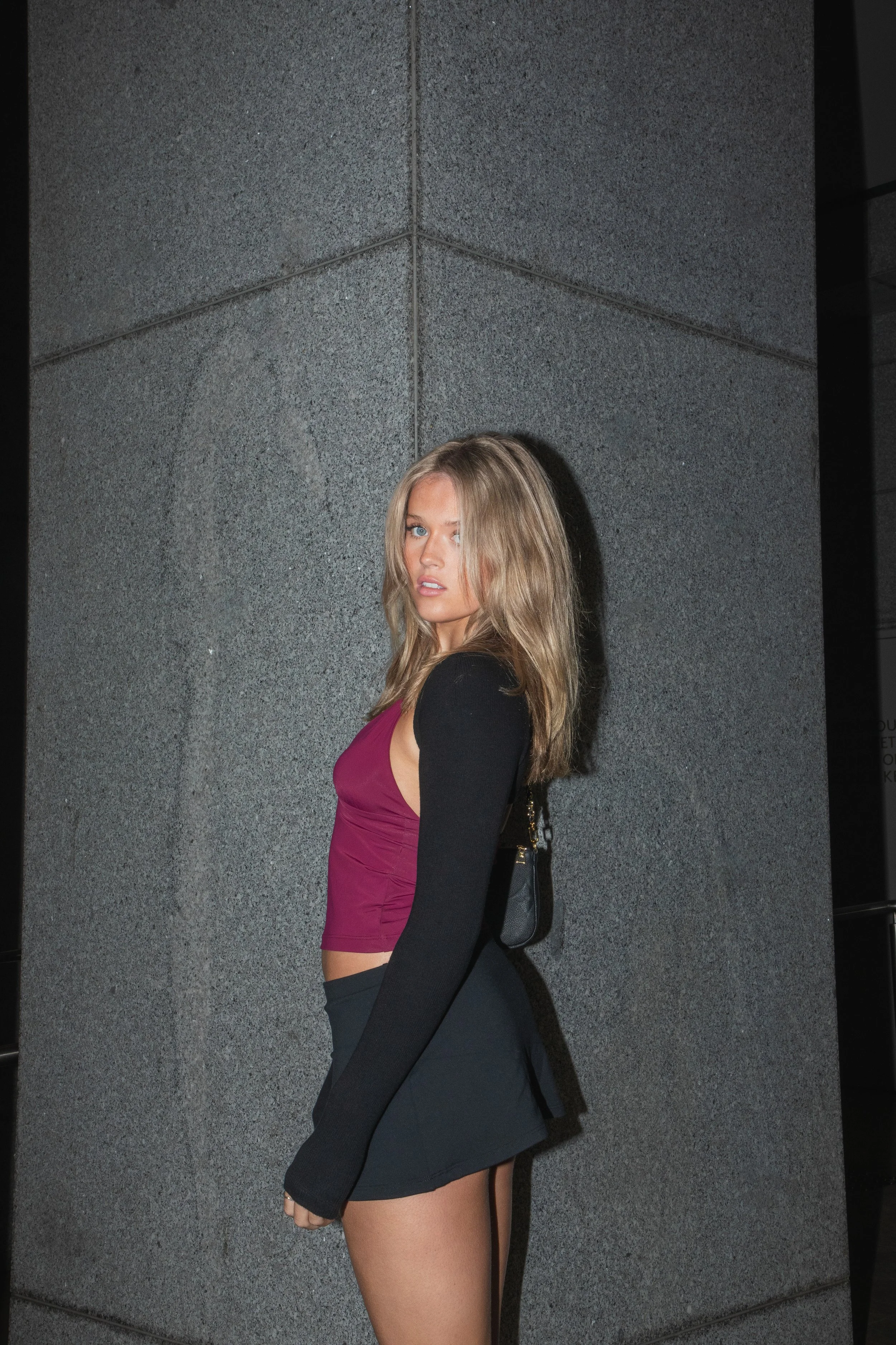 A young woman with long blonde hair and blue eyes stands against a gray concrete wall, wearing a sleeveless burgundy top and a black mini skirt, with a small black purse hanging from her shoulder.