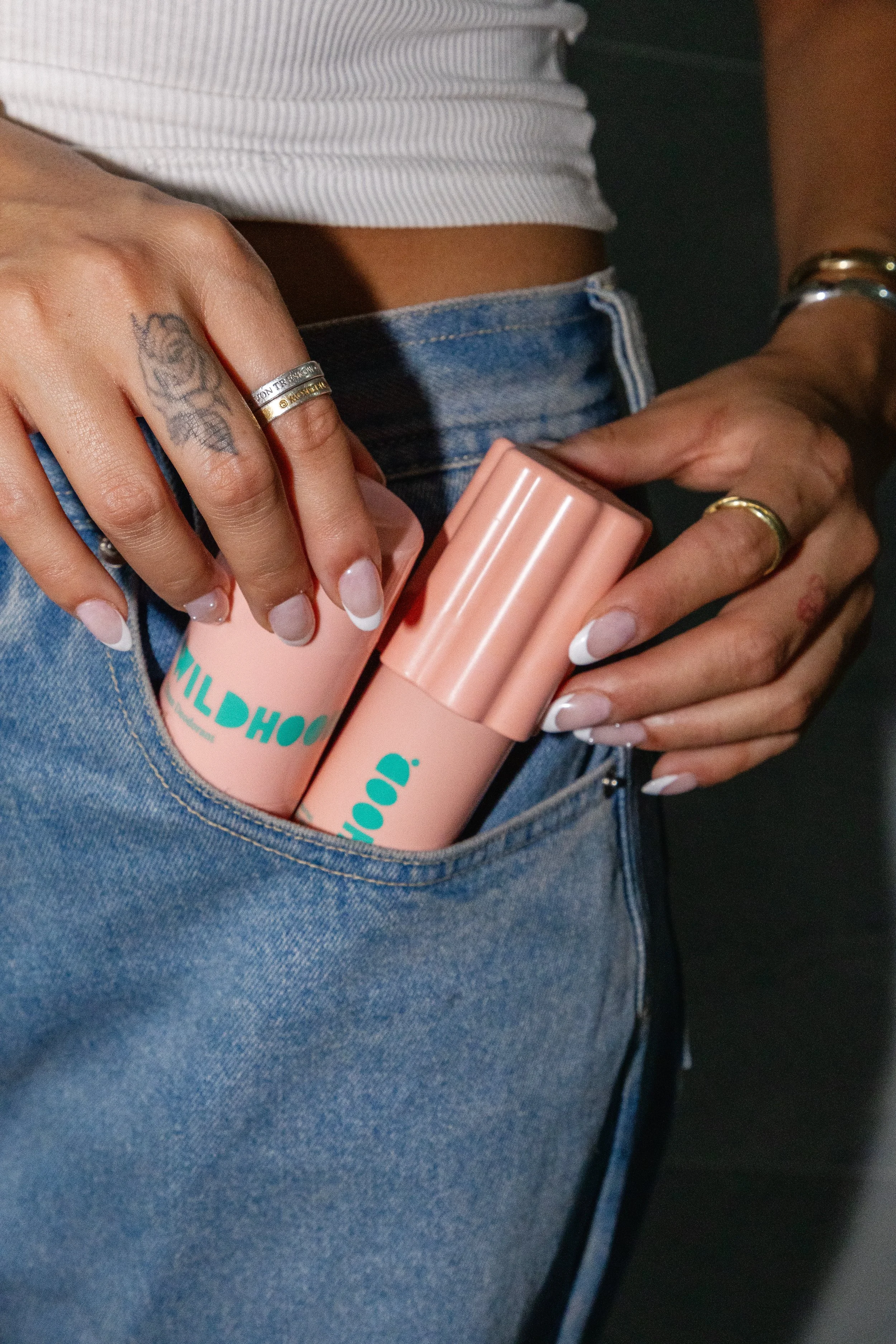 Person with tattoos on fingers, wearing rings and bracelets, placing two pink cylindrical containers with teal text in back pocket of blue jeans.