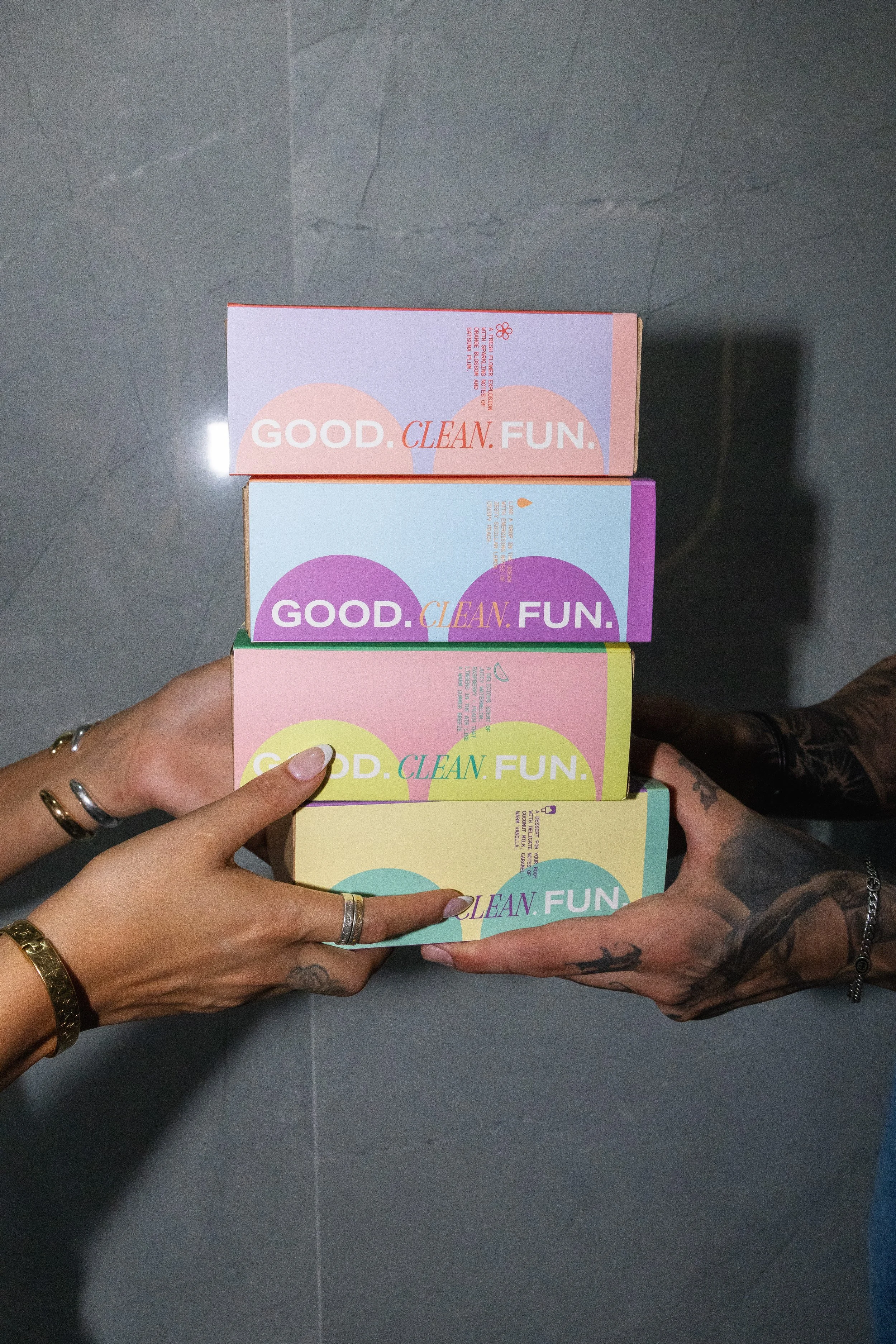 Two hands, one with tattoos and the other with rings, holding a stack of five colorful boxes with the words 'GOOD. CLEAN. FUN.' printed on each. The boxes are pastel pink, purple, yellow, and blue, arranged against a gray tiled background.