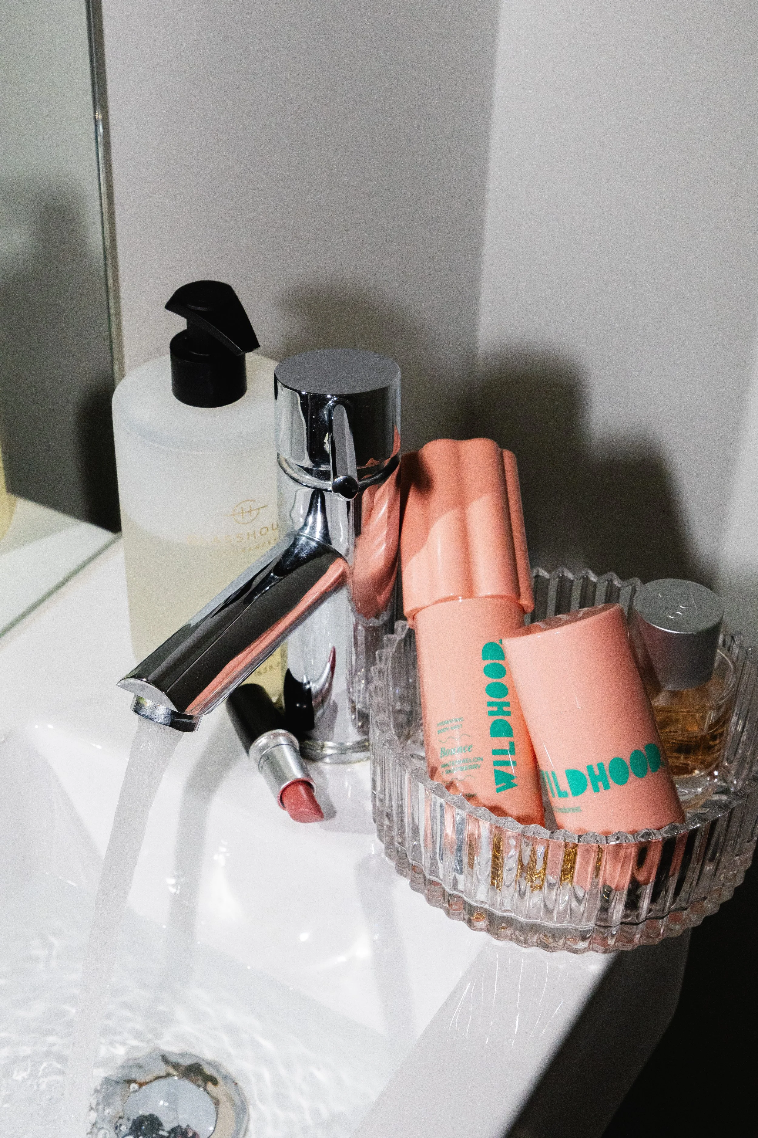 Bathroom sink with water running, a bottle of hand wash, a silver faucet, a lipstick, and a glass dish containing pink and gold bottles