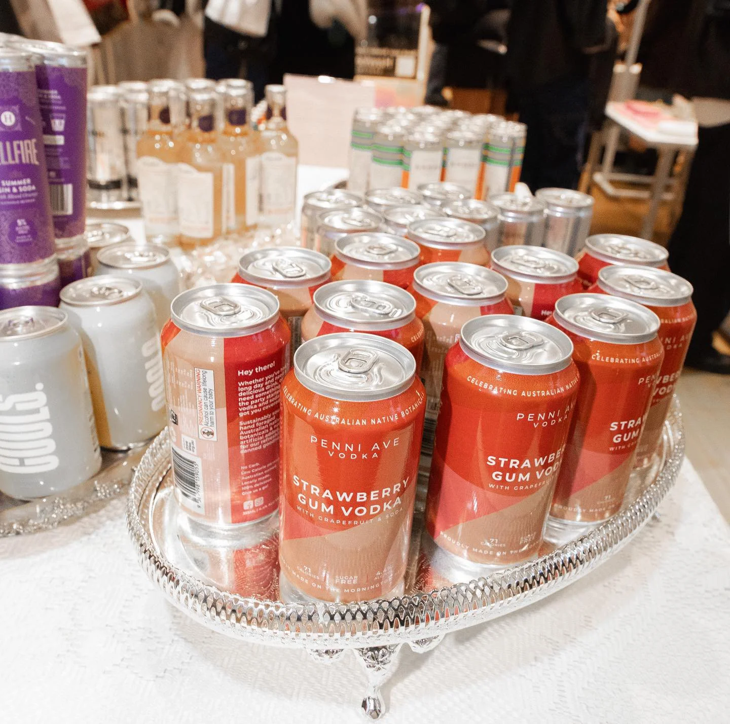 A silver tray with cans of Strawberry Gum Vodka on a white tablecloth, with bottles and other canned drinks in the background.