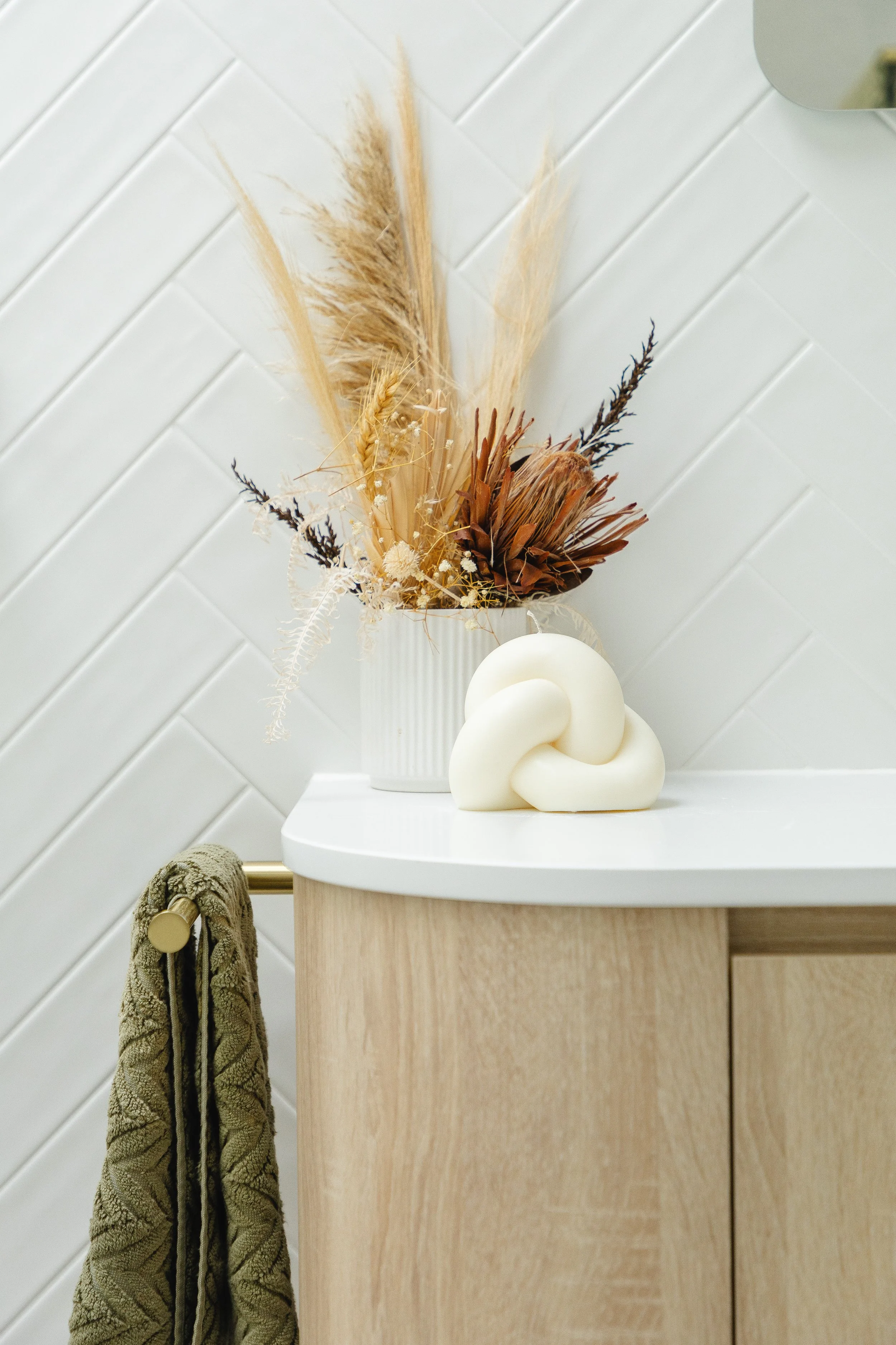 Decorative vase with dried plants and a modern white sculpture on a light wood cabinet, with a green towel hanging on a brass hook.
