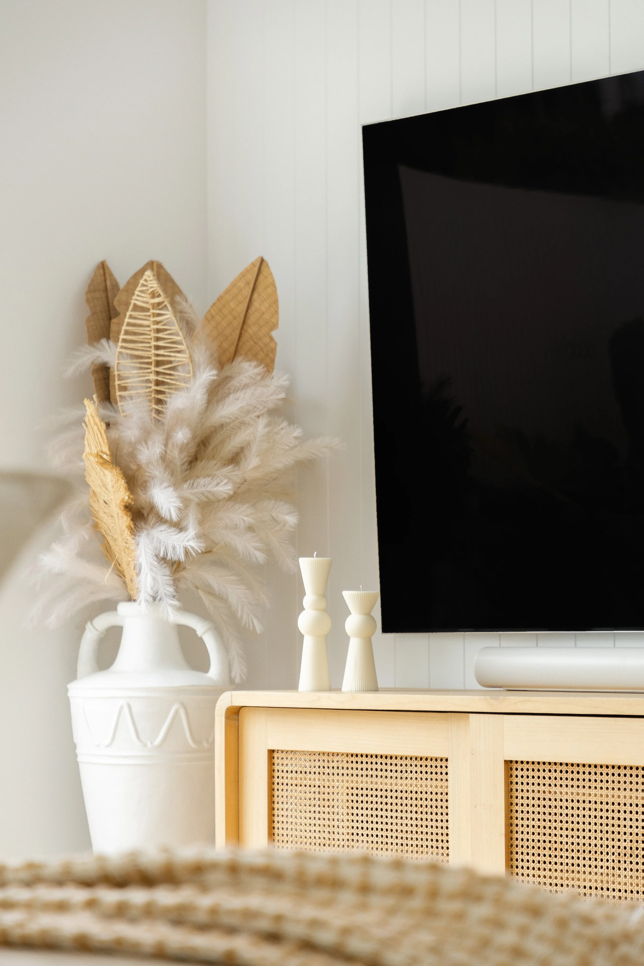 A decorative white vase holding brown paper leaves and white feathers, placed on a light wooden cabinet next to two white candles, and a large black TV on a white wall.