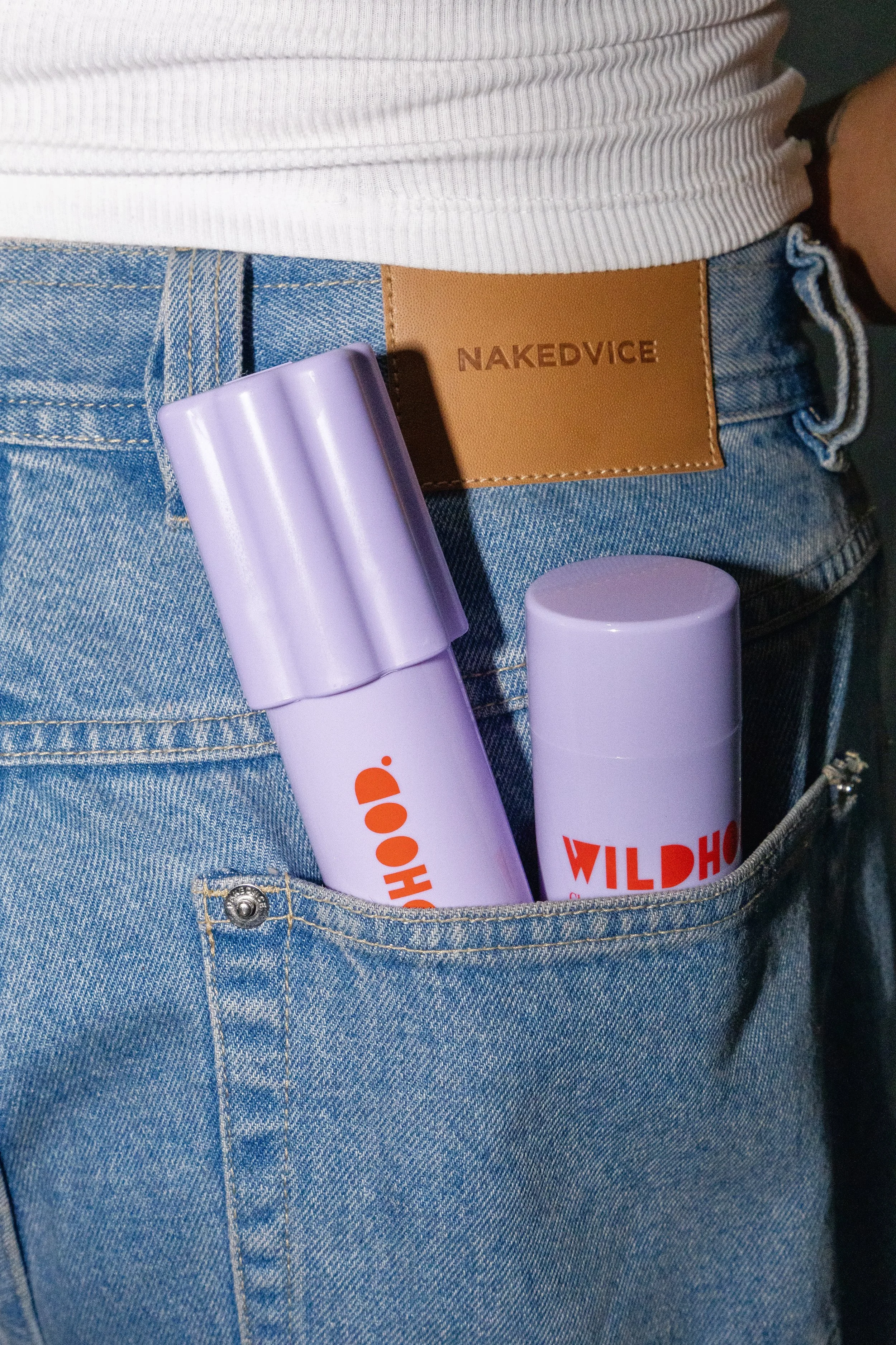 Close-up of a person's front pocket with two purple skincare or cosmetic containers labeled "WILDHO" and "WILDHO" inside, and a tan leather belt with the brand name "NAKEDVICE." they are wearing blue jeans and a white ribbed top.
