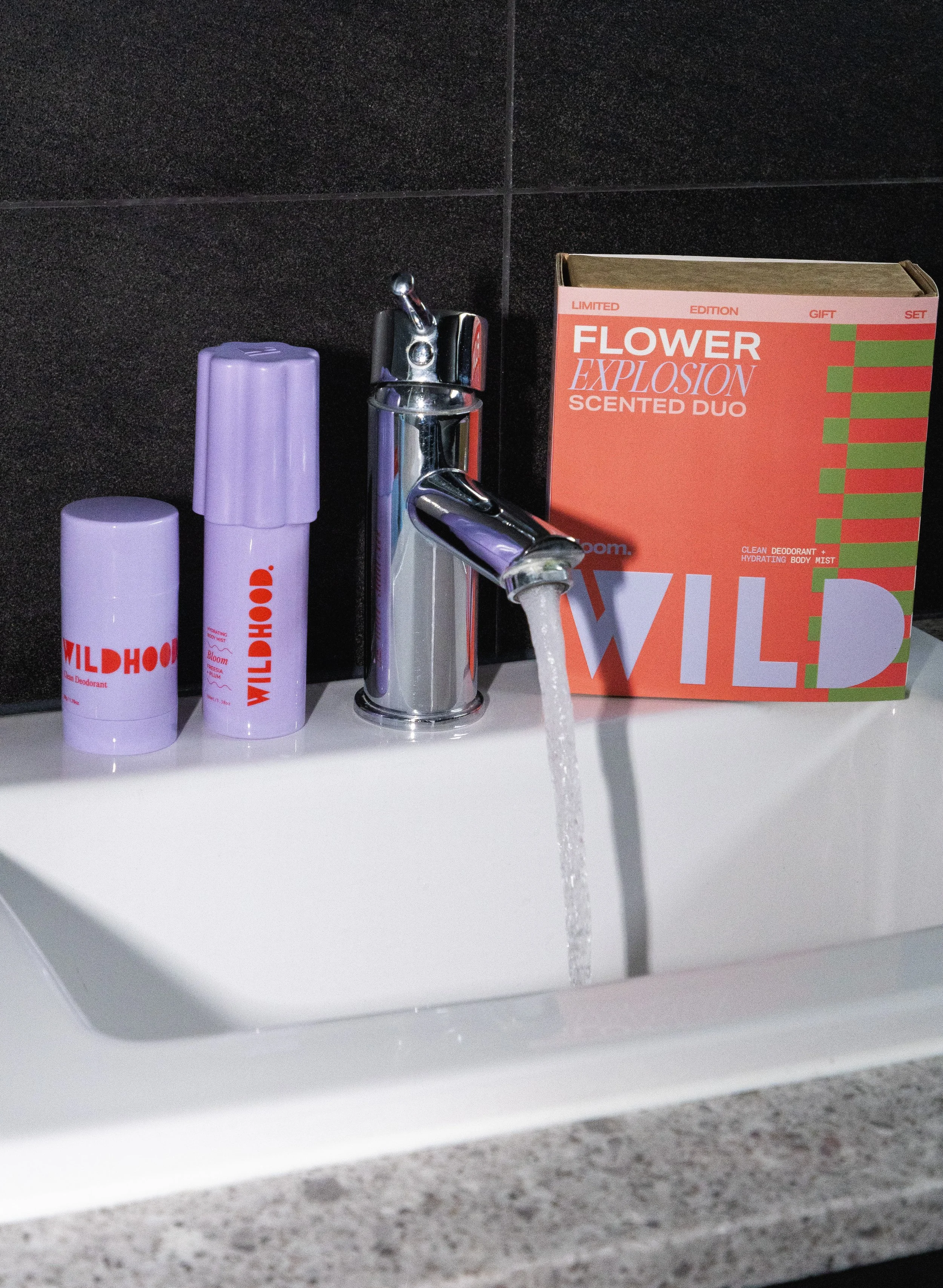 A sink with a chrome faucet running water, two purple Wildhoo deodorant sticks, and a red and green box labeled 'Flower Explosion Scented Duo' on a black tiled wall.