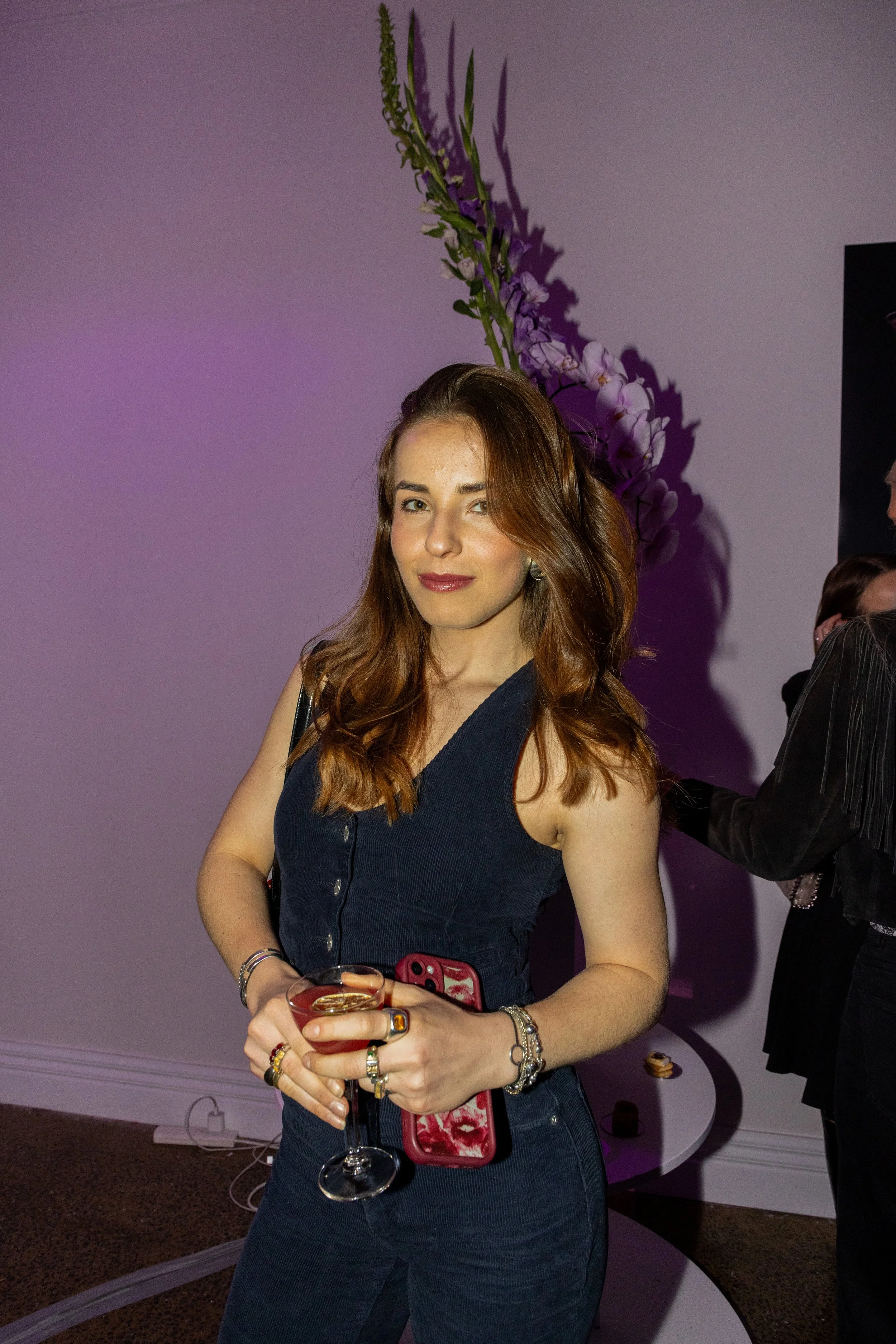A woman with long red hair in a sleeveless dark denim outfit, holding a glass of a pink cocktail, standing in front of a purple-lit wall behind a tall floral arrangement with purple and white flowers, at an indoor social event.