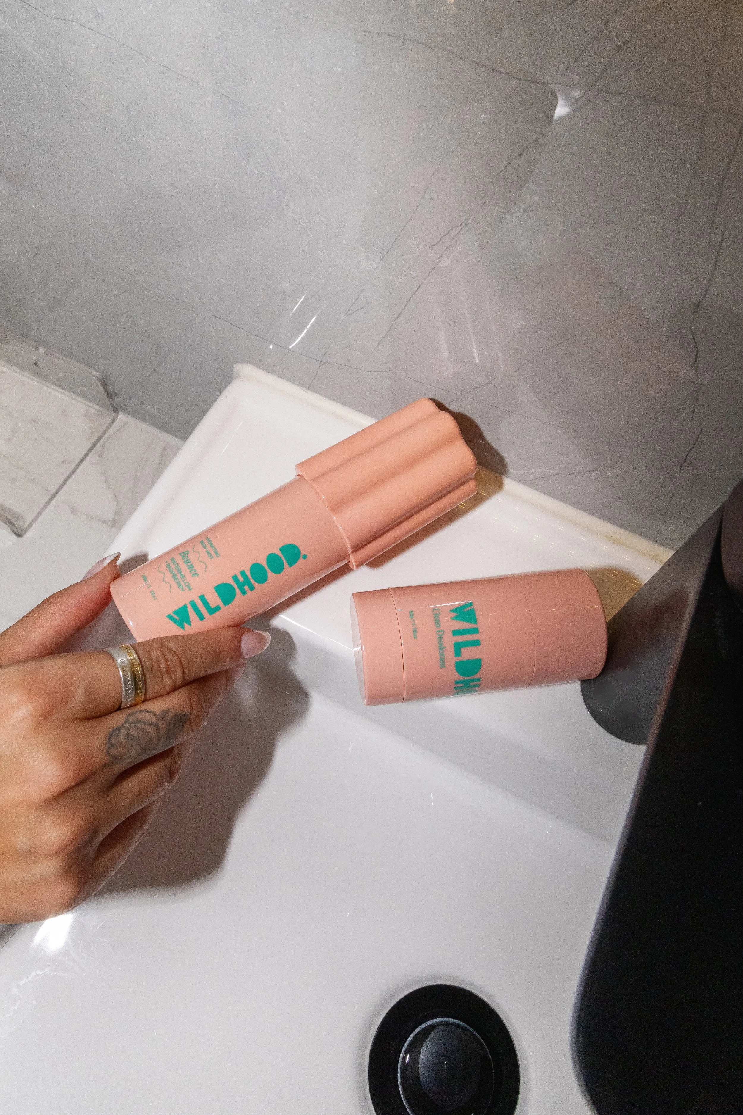 A hand with a ring and a tattoo holding a pink cylindrical container labeled 'WILDHOOD' beside a pink tube with the same branding, on a white surface in front of a gray tiled wall.