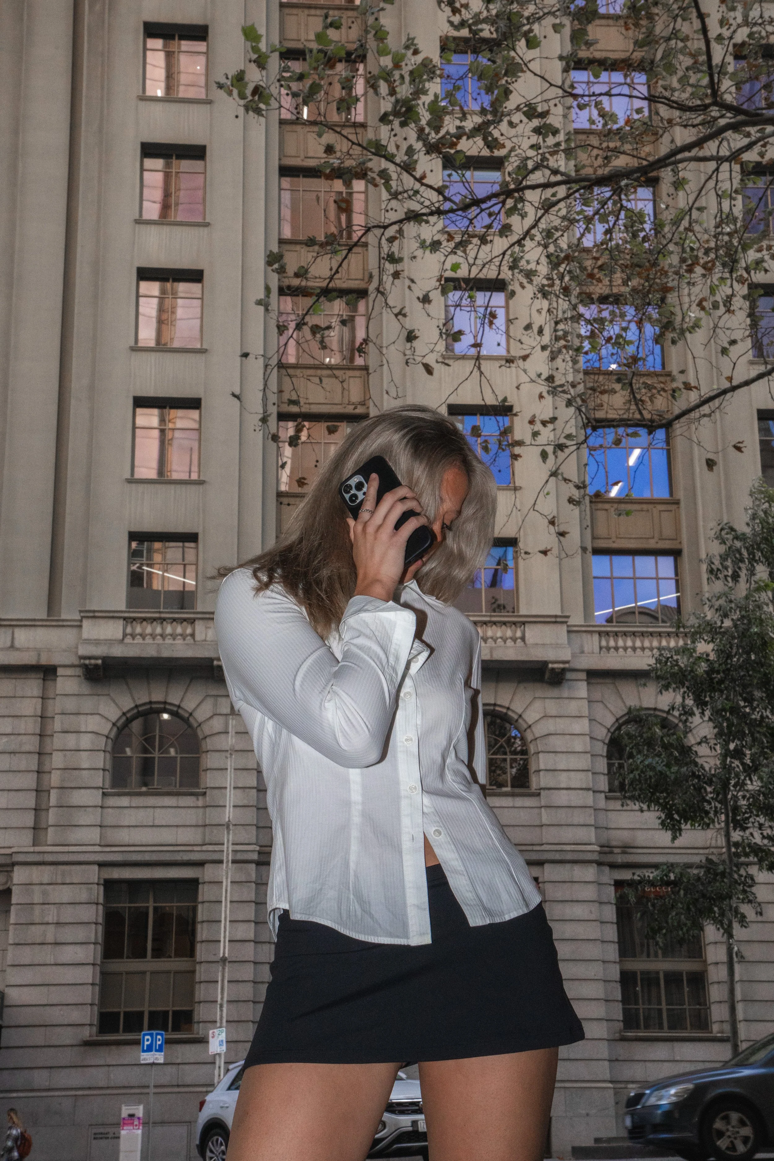 A woman with blonde hair, wearing a white button-up shirt and black skirt, talking on a mobile phone outside in an urban area with tall buildings and parked cars, during dusk or evening.