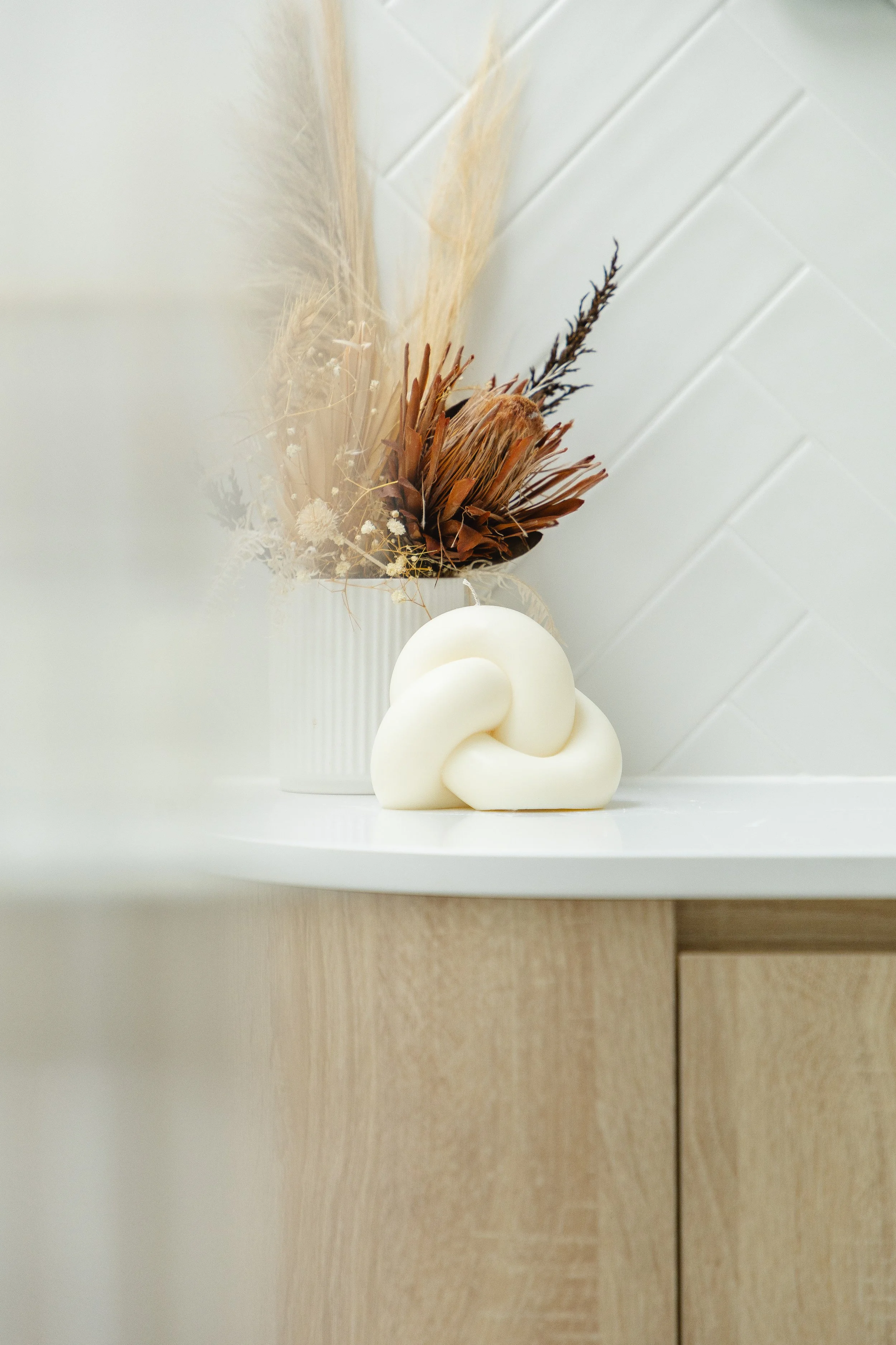 Decorative arrangement with dried flowers and a white elastic knot sculpture on a white surface against a white tiled wall.