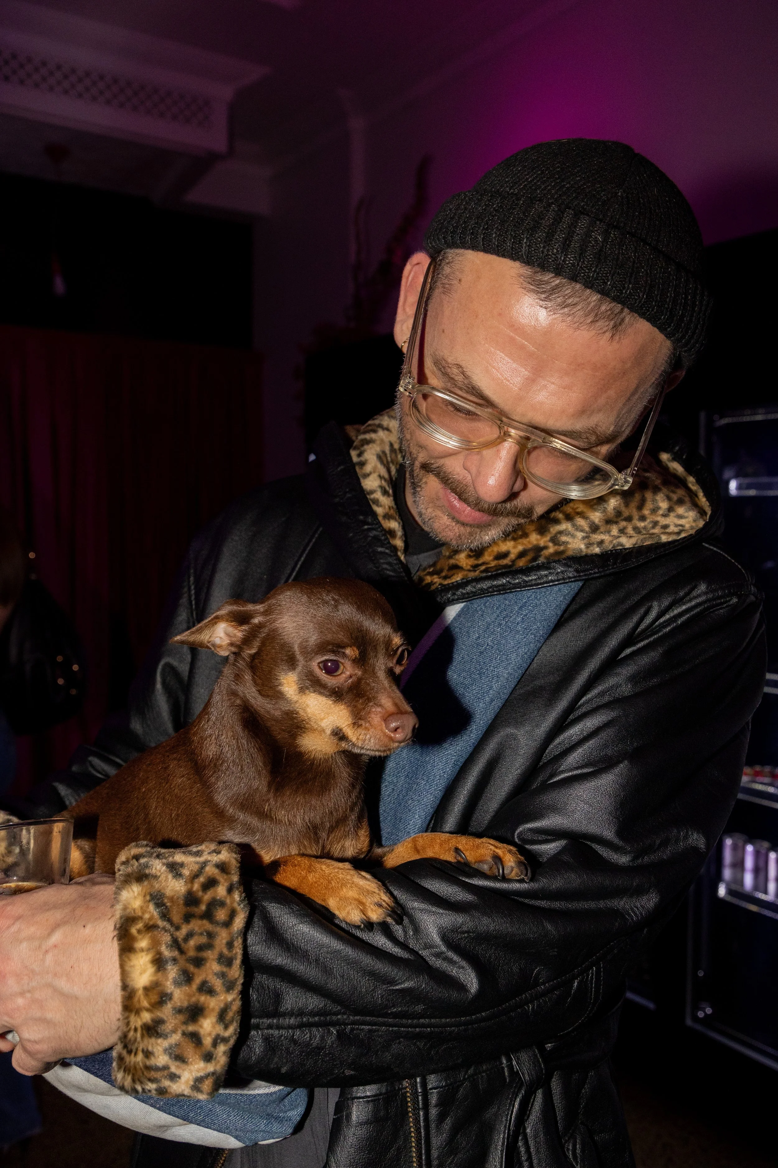 A man wearing glasses, a black beanie, a black leather jacket with leopard print lining, is holding a small brown dog with tan markings. The man is looking down at the dog, which is resting in his arms.