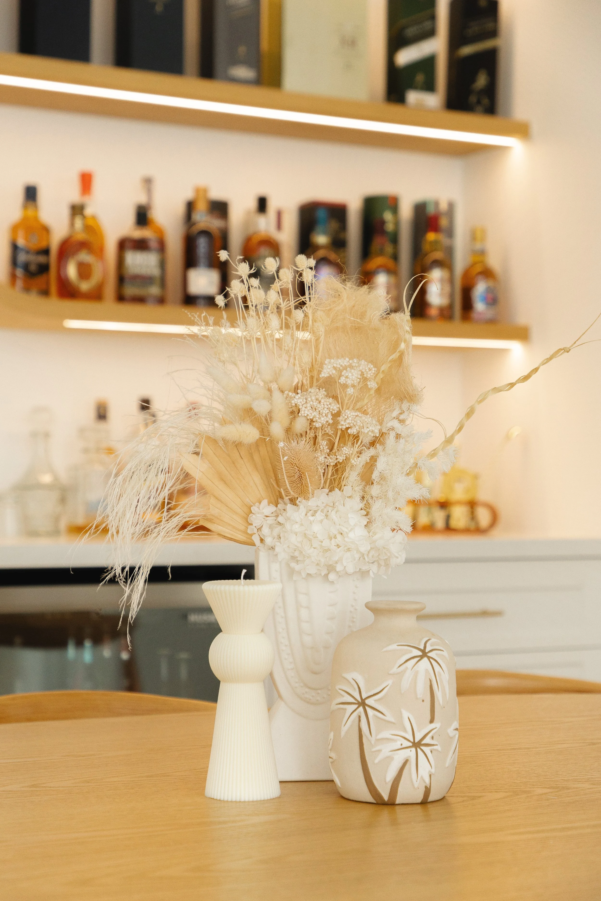 Decorative dried flower arrangement in a white vase, with a smaller cream-colored vase and a beige vase with tropical leaf design on a wooden table.