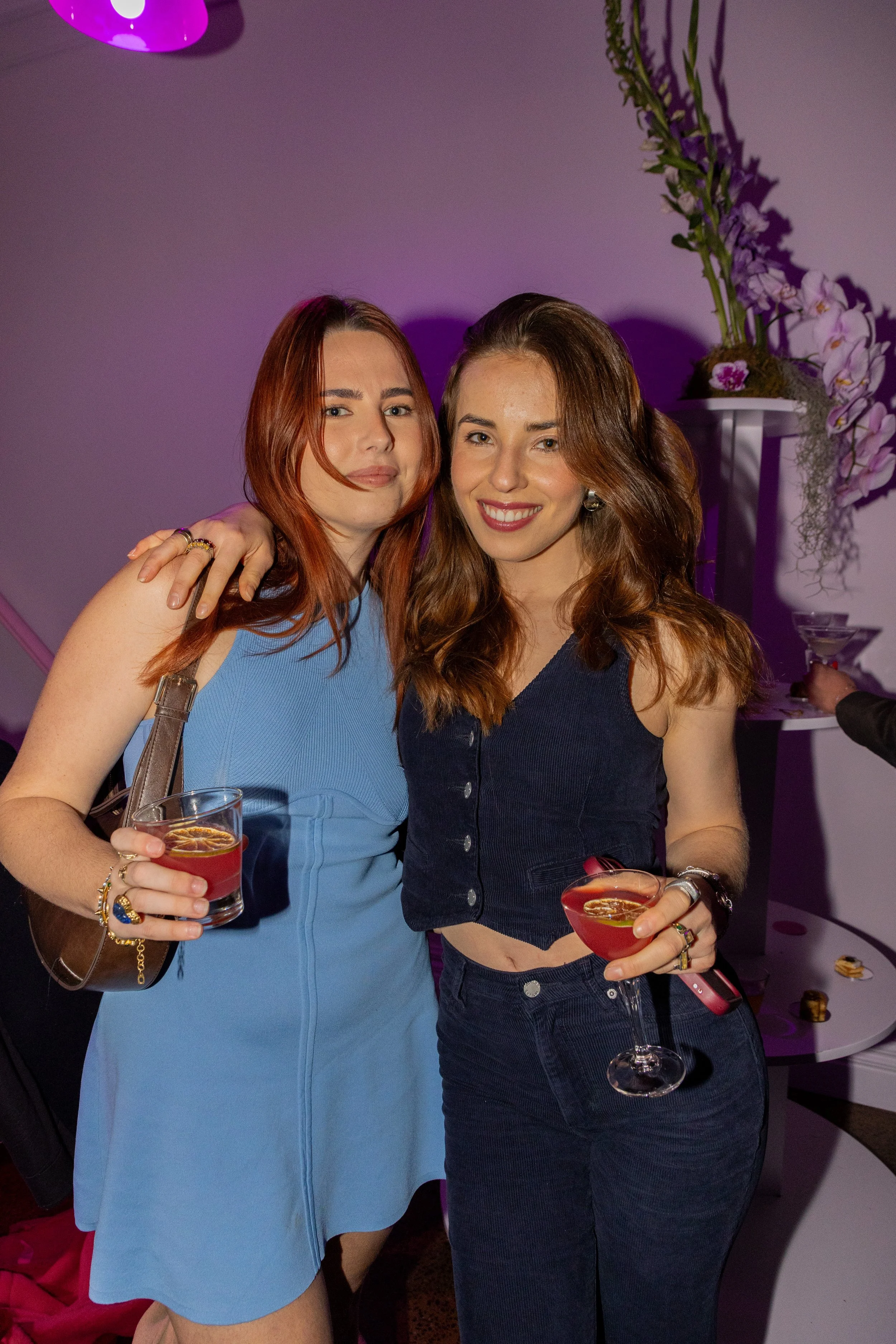 Two young women at a party, smiling and holding cocktails, with purple lighting in the background and floral decorations nearby.