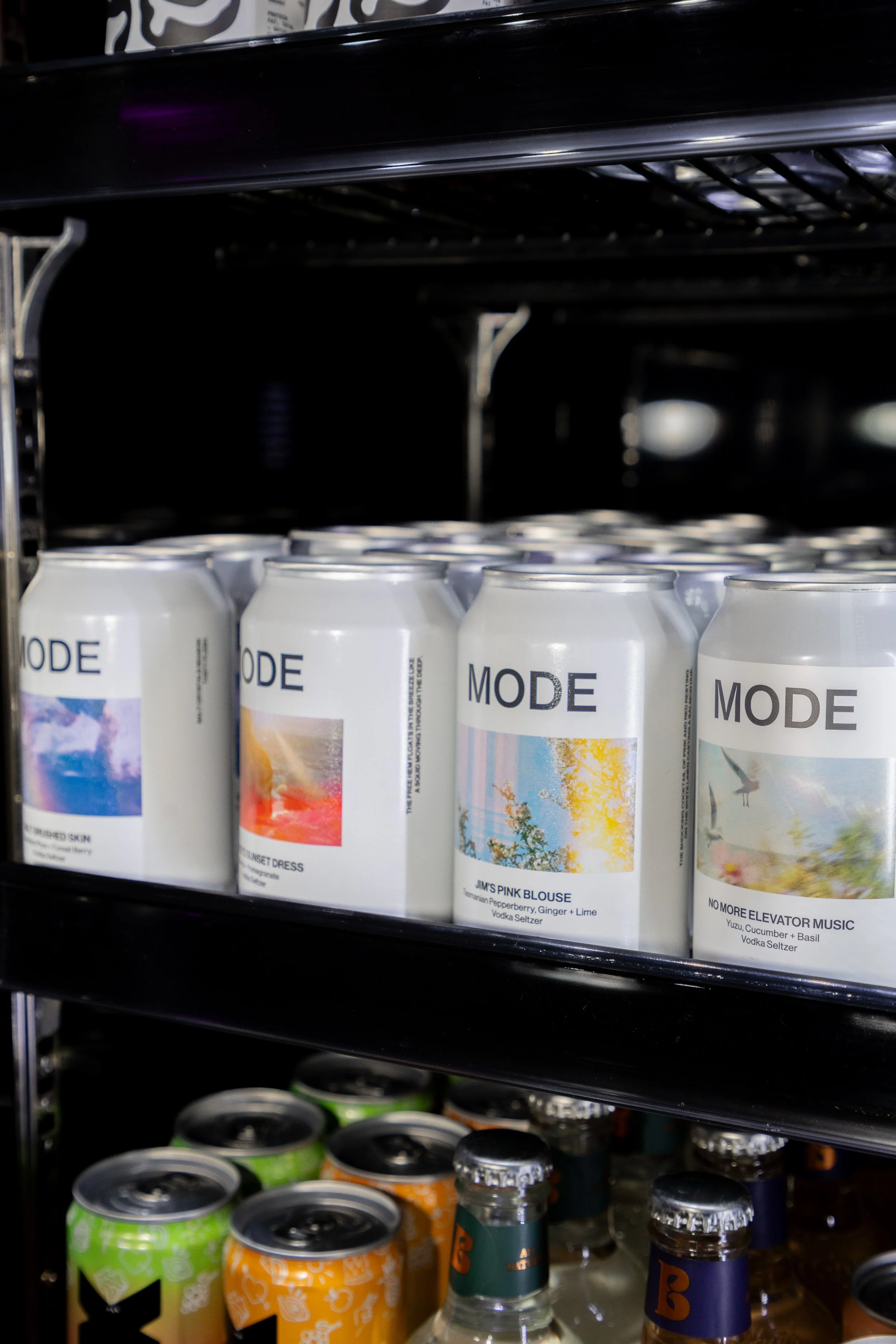 Refrigerator shelf displaying cans of MODE beverage, each with colorful abstract labels, with smaller cans and bottles underneath.