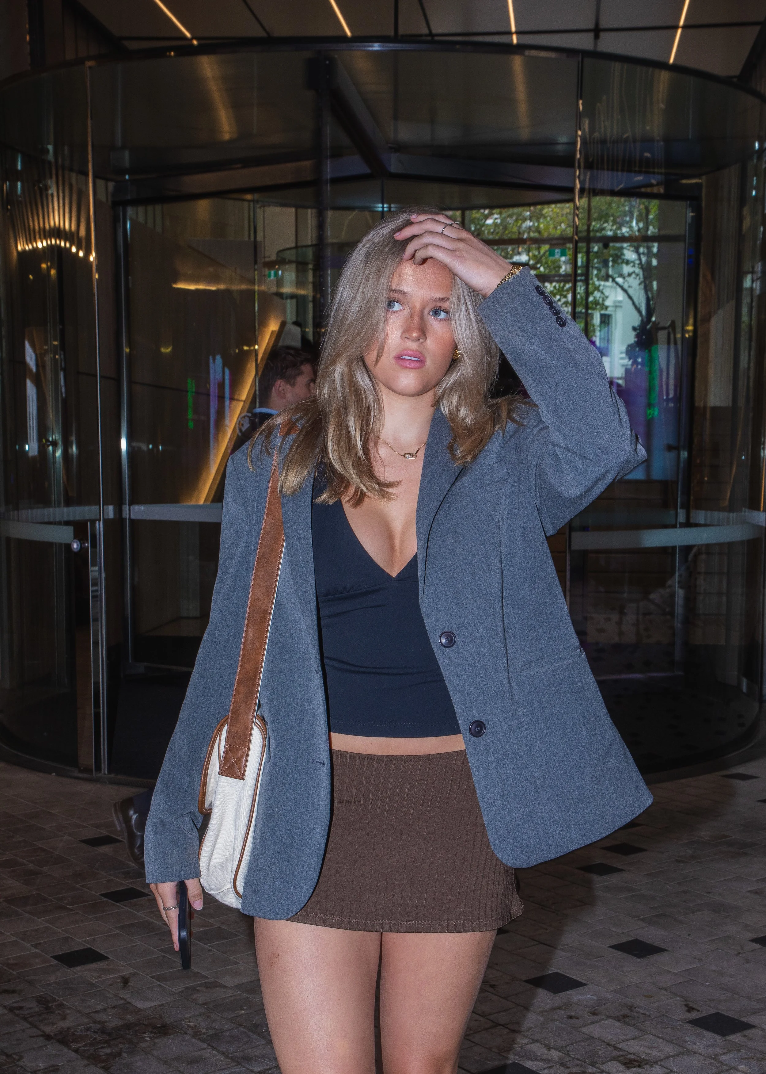 A young woman with wavy blonde hair is standing outside in front of a revolving door. She is wearing a gray blazer, a black crop top, and a brown mini skirt. She has a beige shoulder bag and appears to be adjusting her hair with her right hand, looki